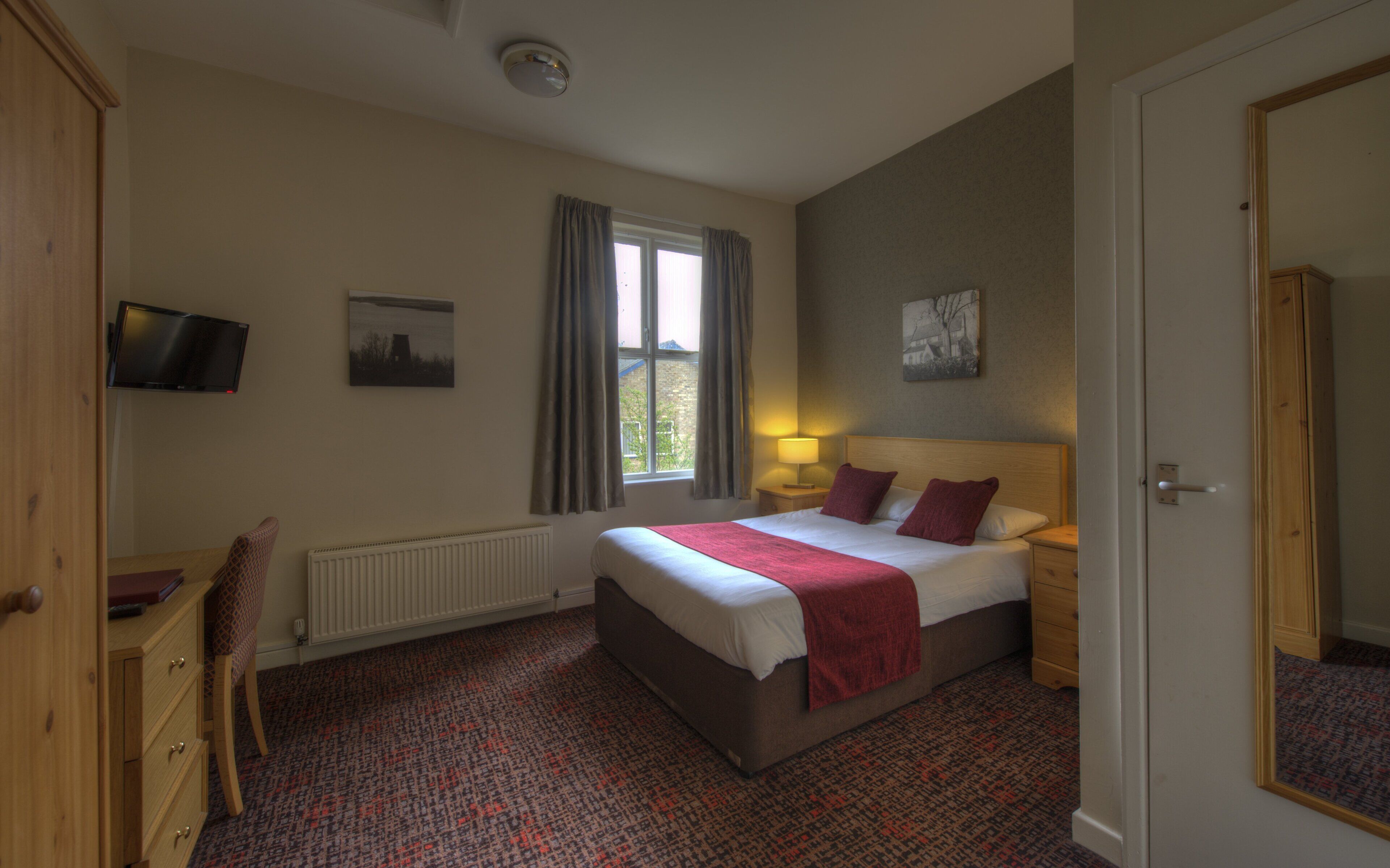 A hotel room with a bed, a desk, a chair, a radiator, a window with curtains, a lamp, and artwork on the walls.