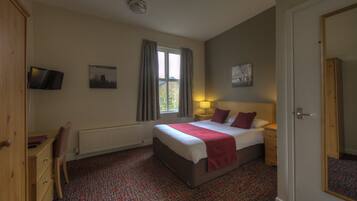 A hotel room with a bed, a desk, a chair, a radiator, a window with curtains, a lamp, and artwork on the walls.