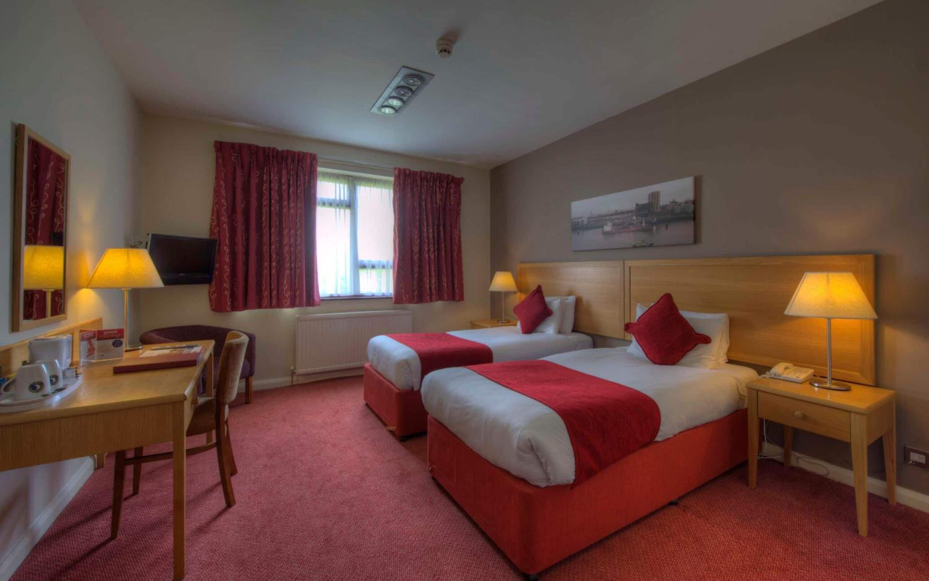 A hotel room with two beds, a desk, a chair, a TV, and a window with curtains.