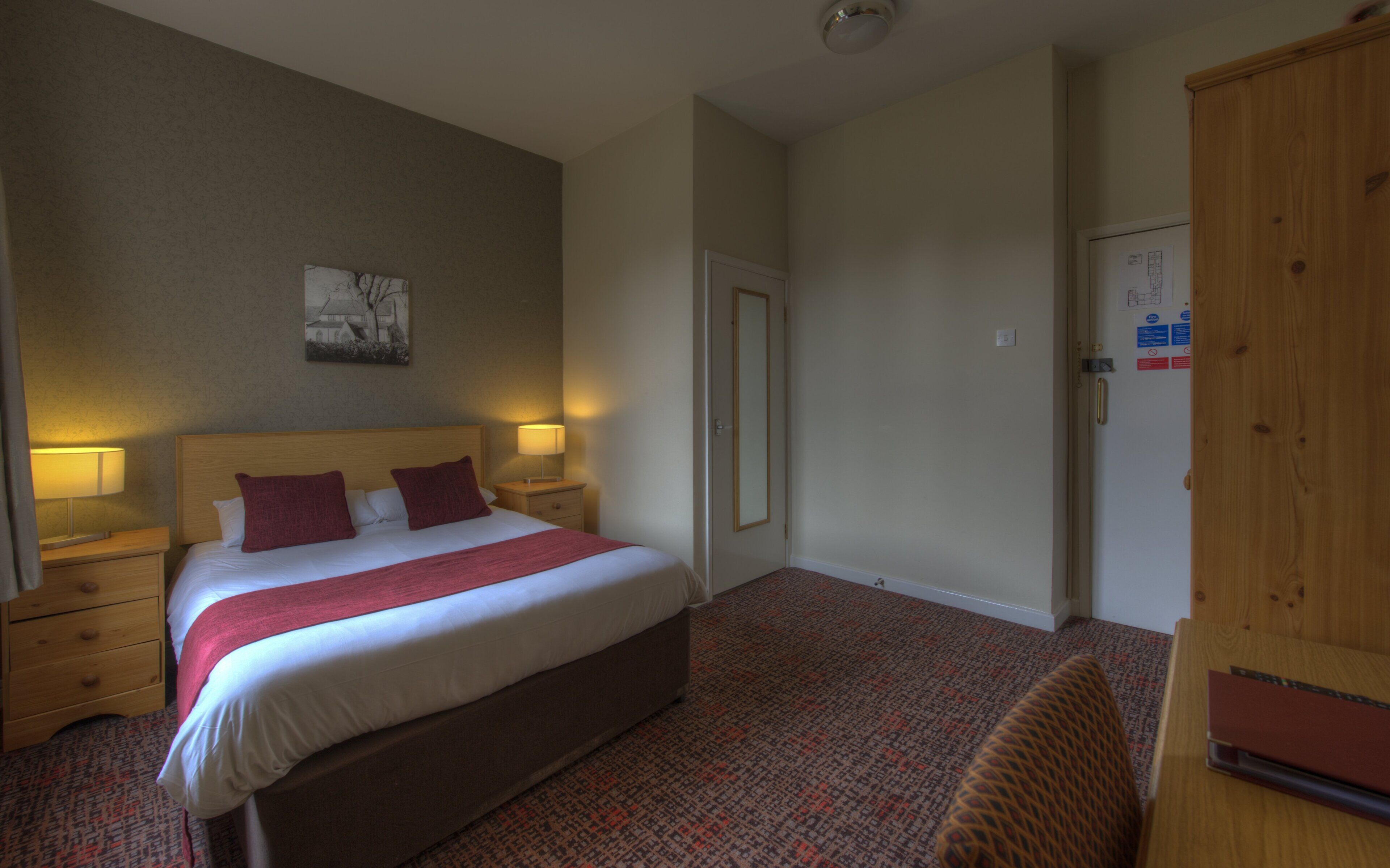 Una habitación de hotel con una cama, dos lámparas de noche, una mesita de noche, un armario de madera y piso alfombrado.