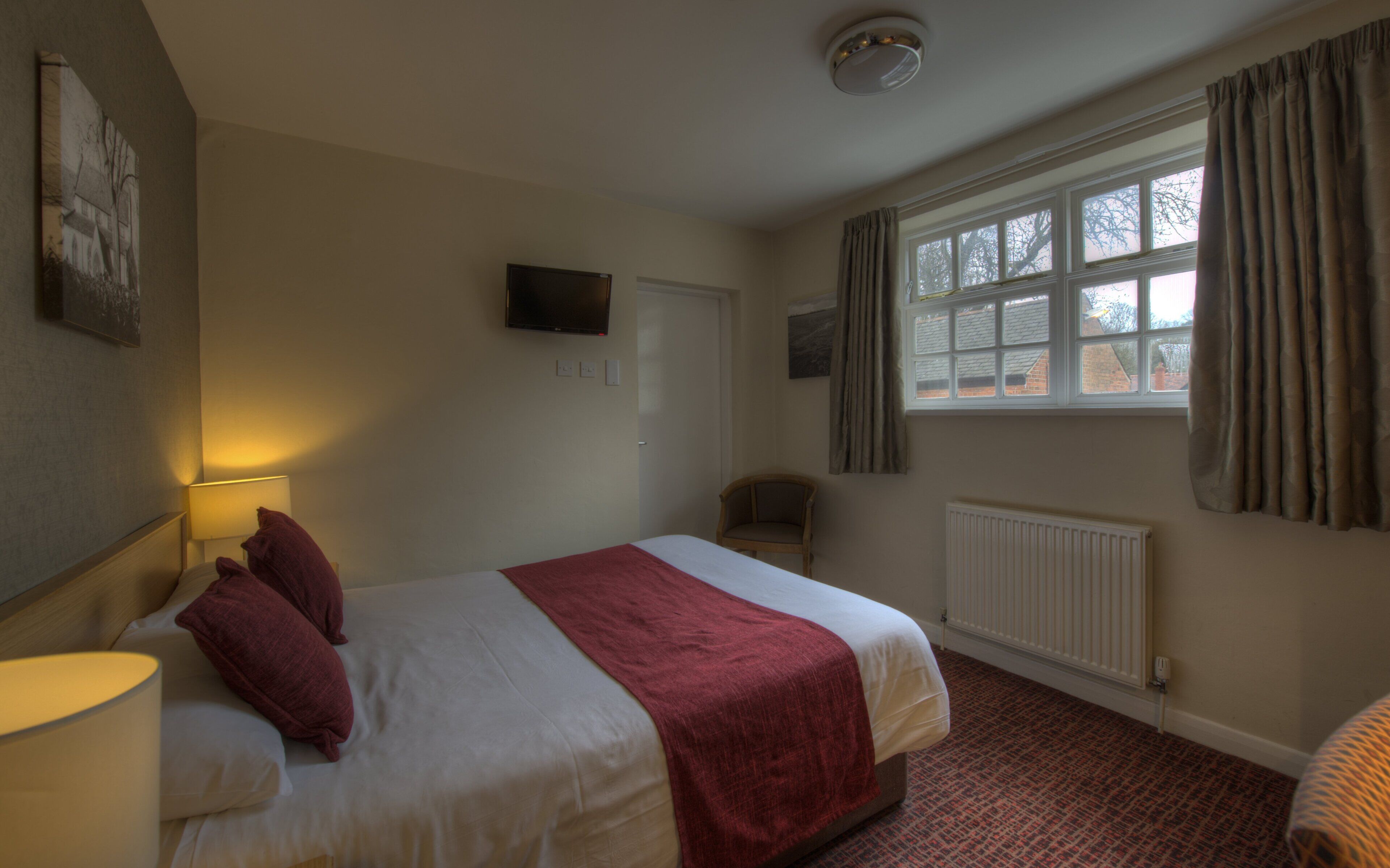 A hotel room with a bed, a chair, a television, a window with curtains, and a radiator.