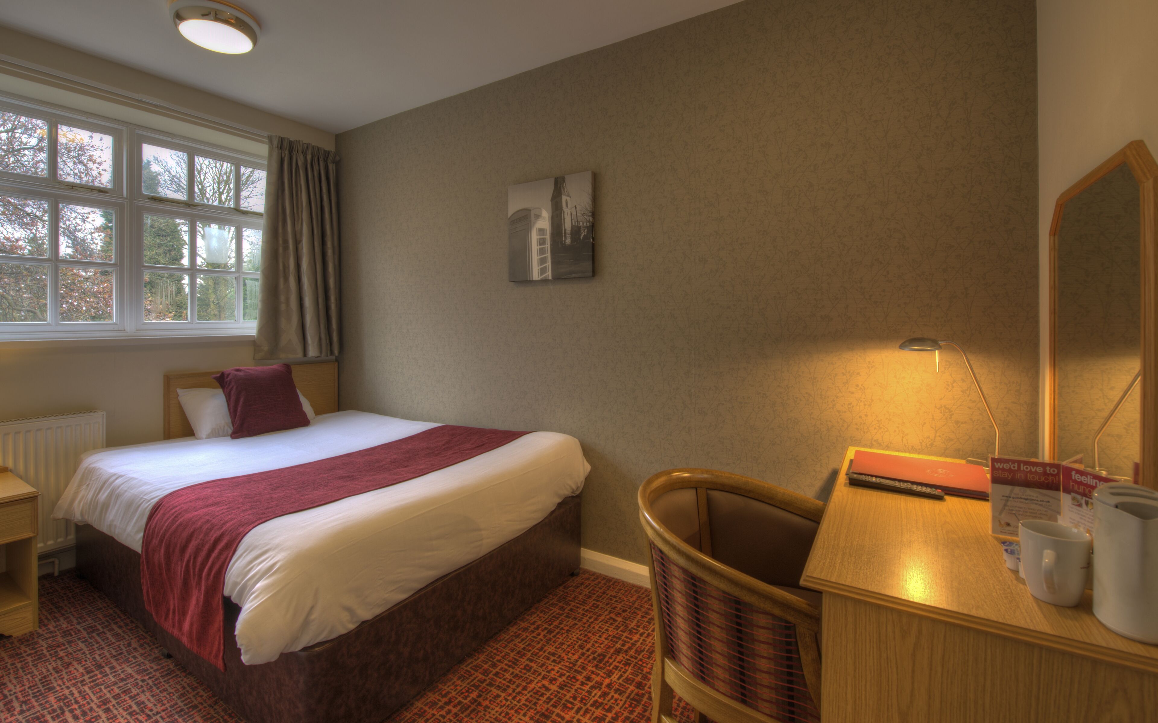 A hotel room with a bed, a desk, a chair, a radiator, a window with curtains, and a wall-mounted picture.