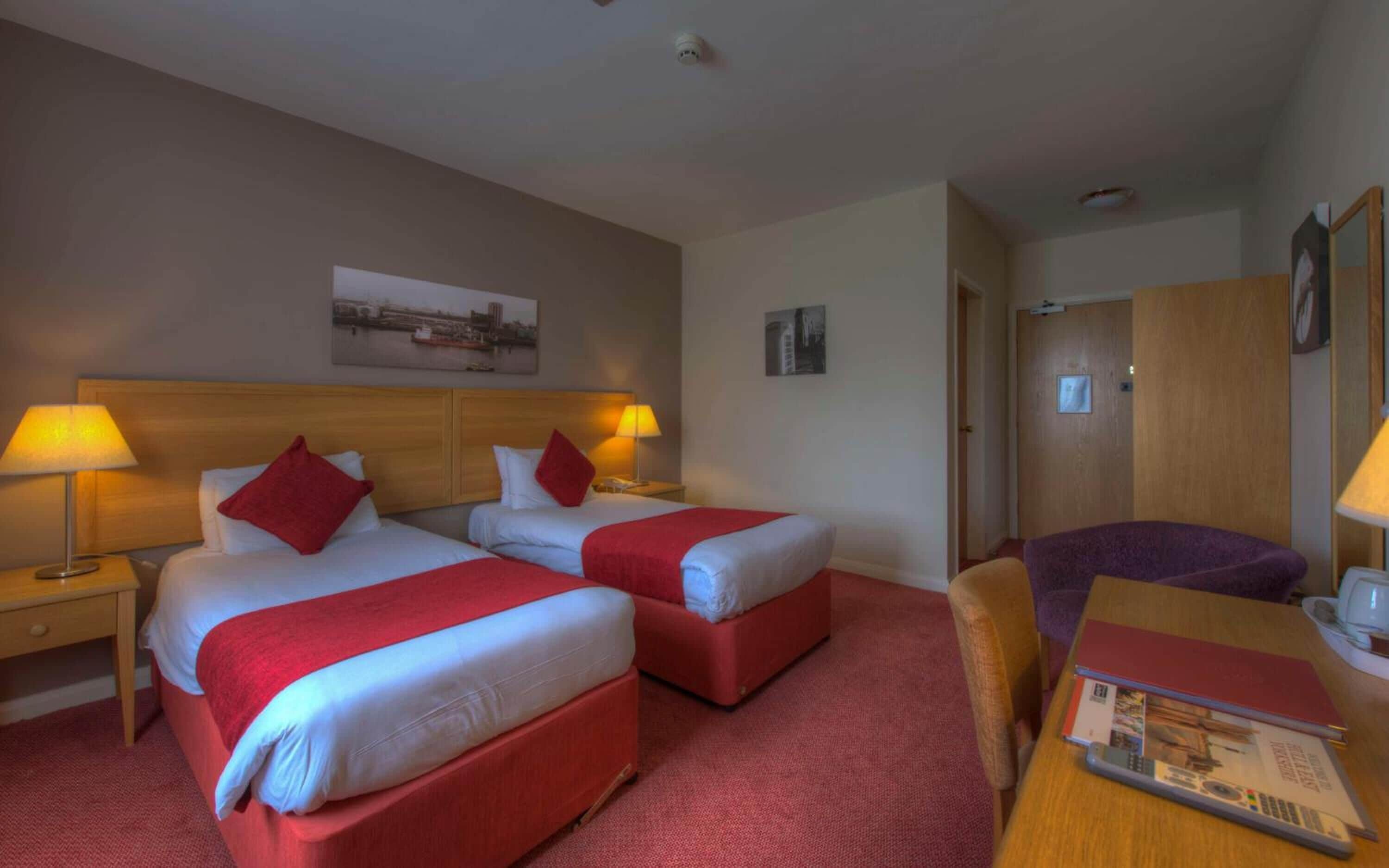 A hotel room with two beds, a desk, a chair, and a door.
