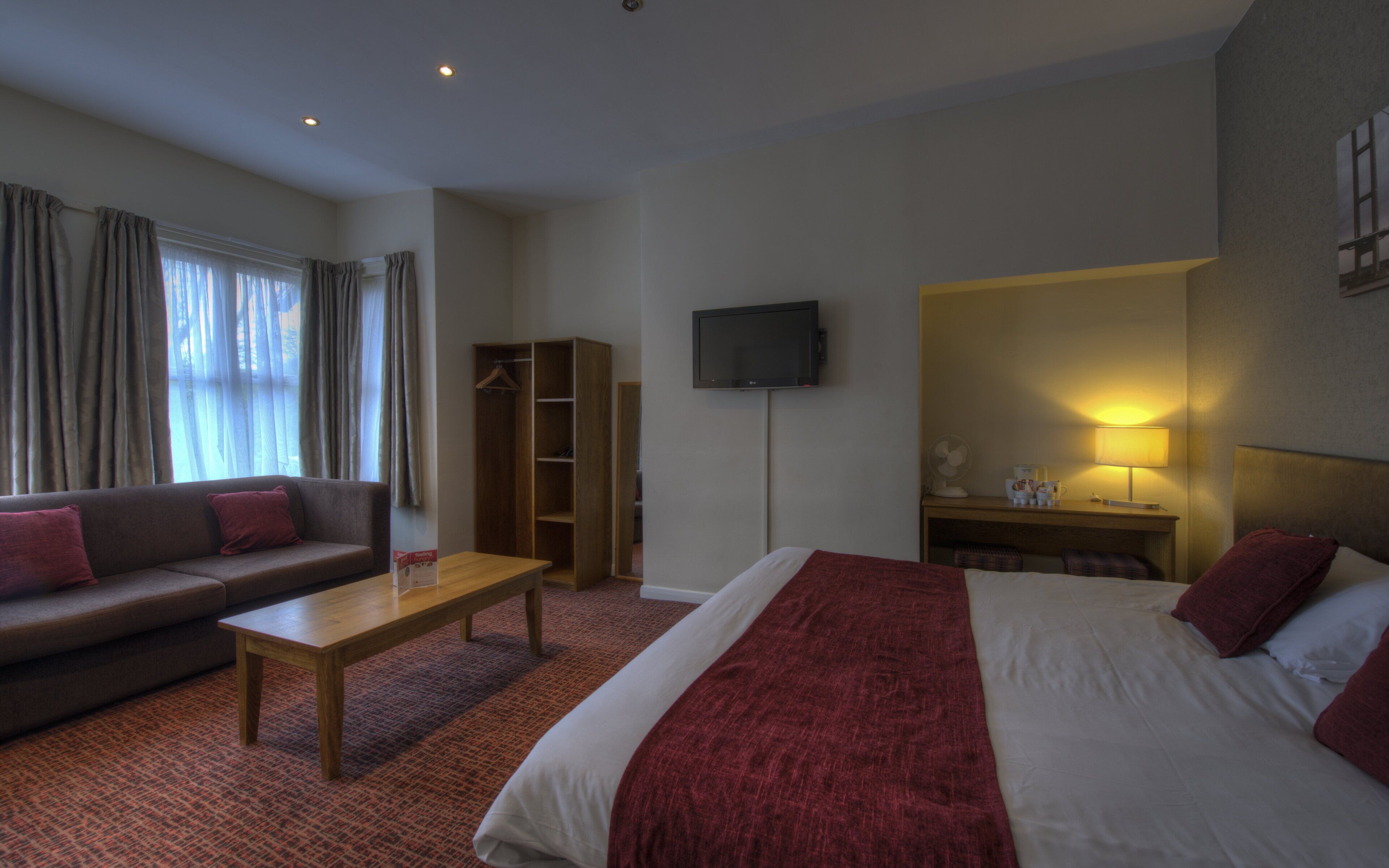 A hotel room with a bed, a sofa, a wooden table, a TV, and a desk with a lamp.