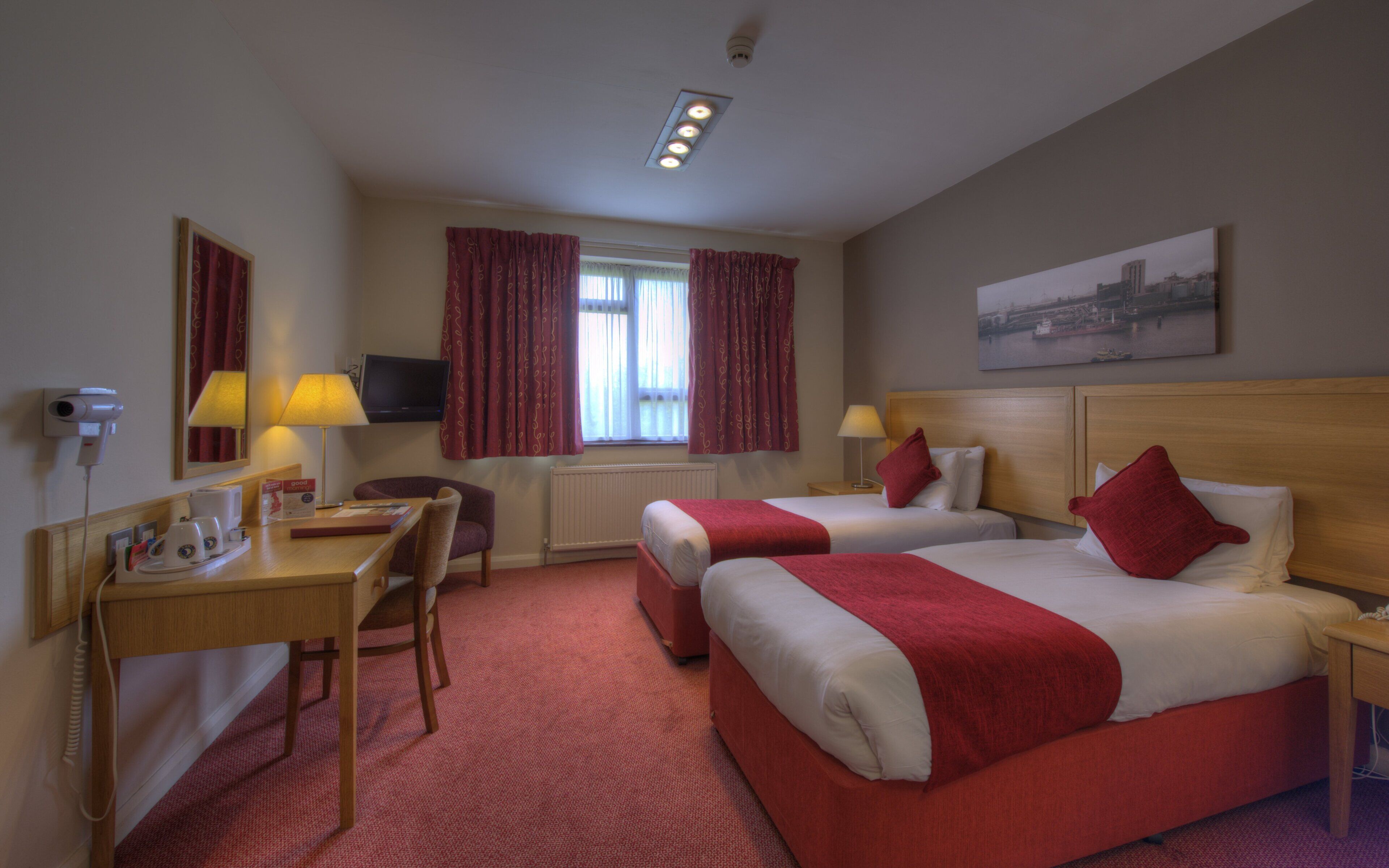 Ein Hotelzimmer mit zwei Betten, einem Schreibtisch, einem Sessel, einem Fernseher und einem Fenster mit Vorhängen.