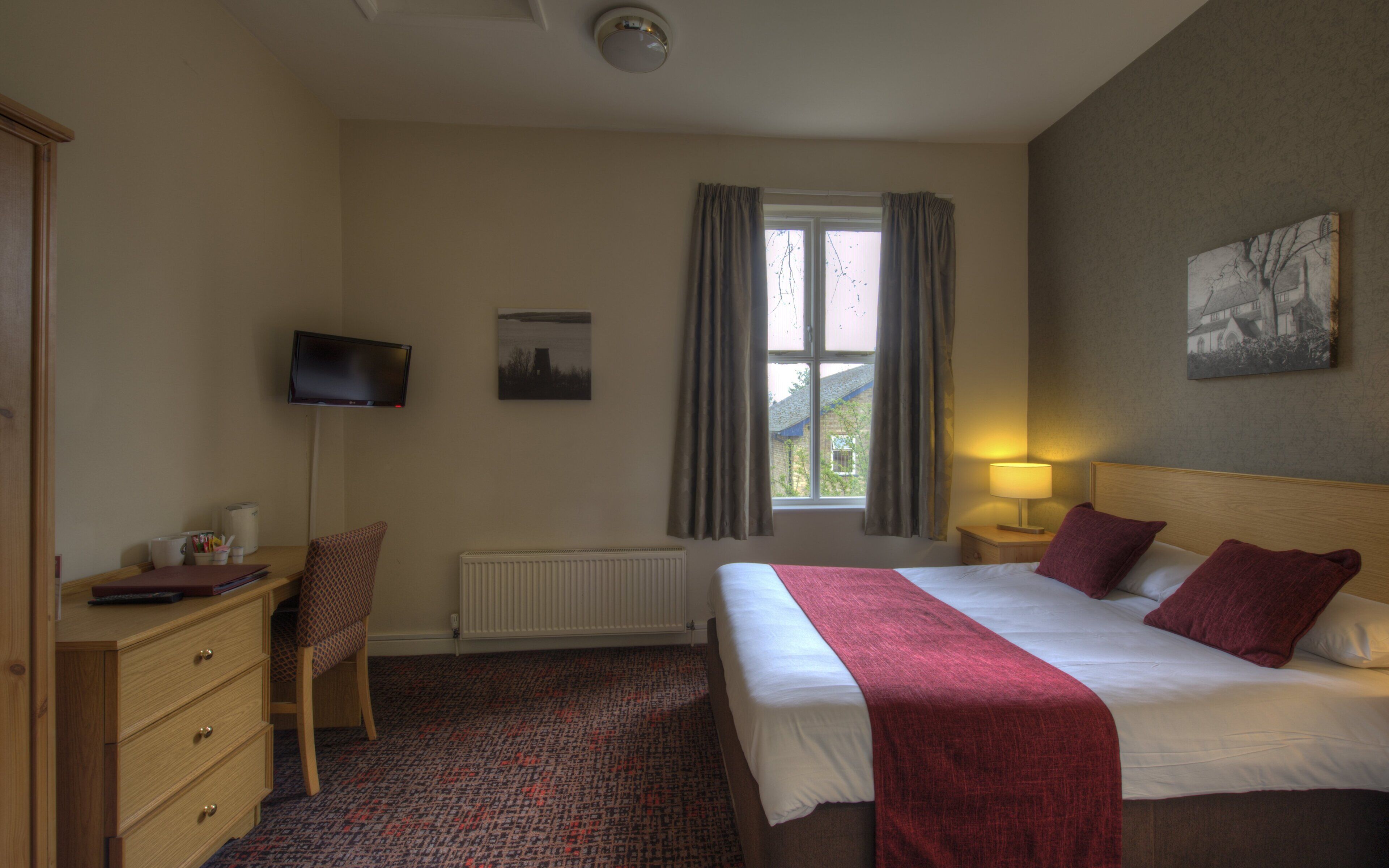 Una camera d'albergo con un letto, una scrivania, una sedia, una televisione, una finestra con tende e un quadro appeso al muro.