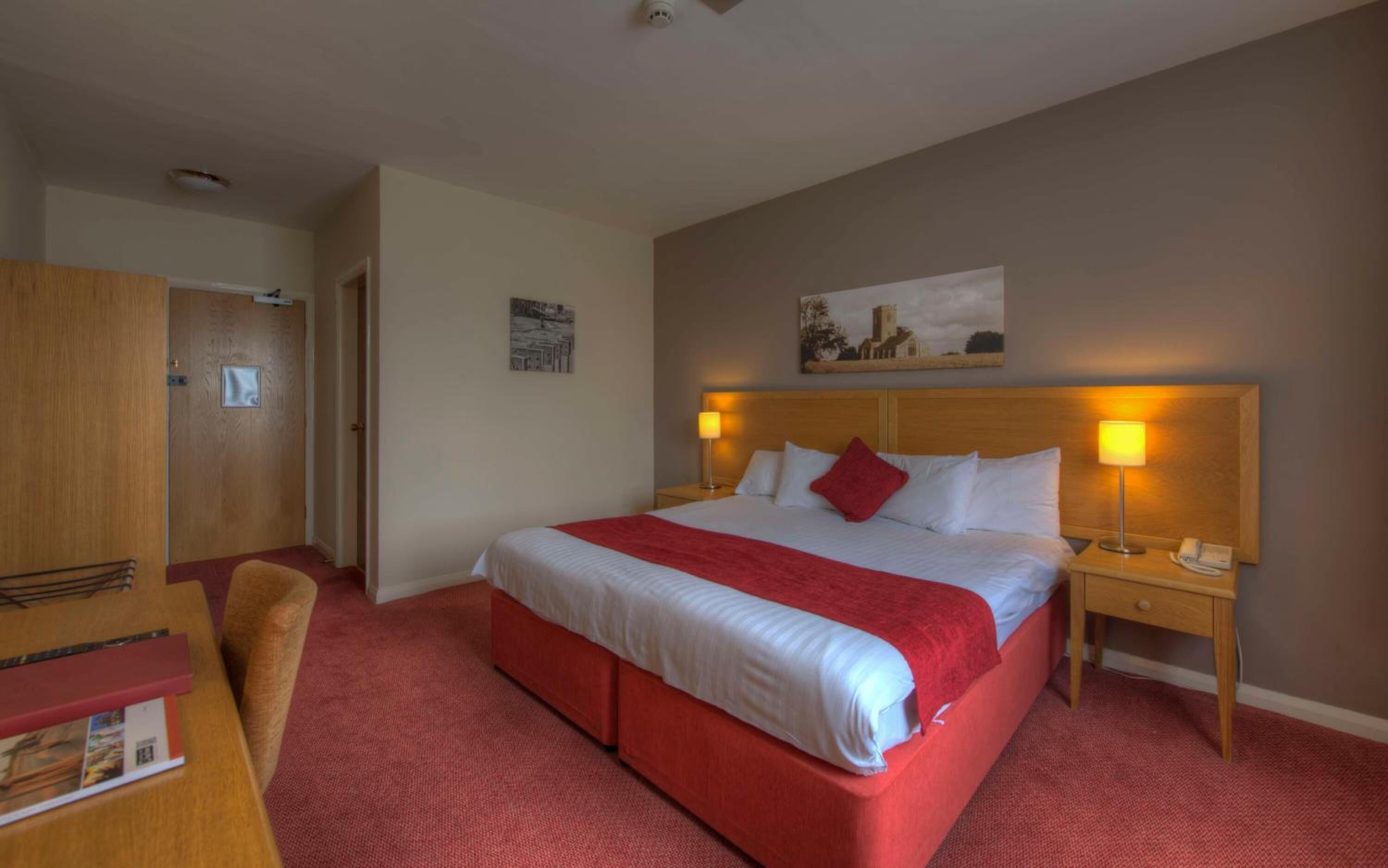 Habitación de hotel con una cama grande, alfombra roja, cabecera de madera, dos lámparas de noche, una mesita de noche, un escritorio de madera con una silla y un cuadro en la pared.