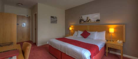 Habitación de hotel con una cama grande, alfombra roja, cabecera de madera, dos lámparas de noche, una mesita de noche, un escritorio de madera con una silla y un cuadro en la pared.