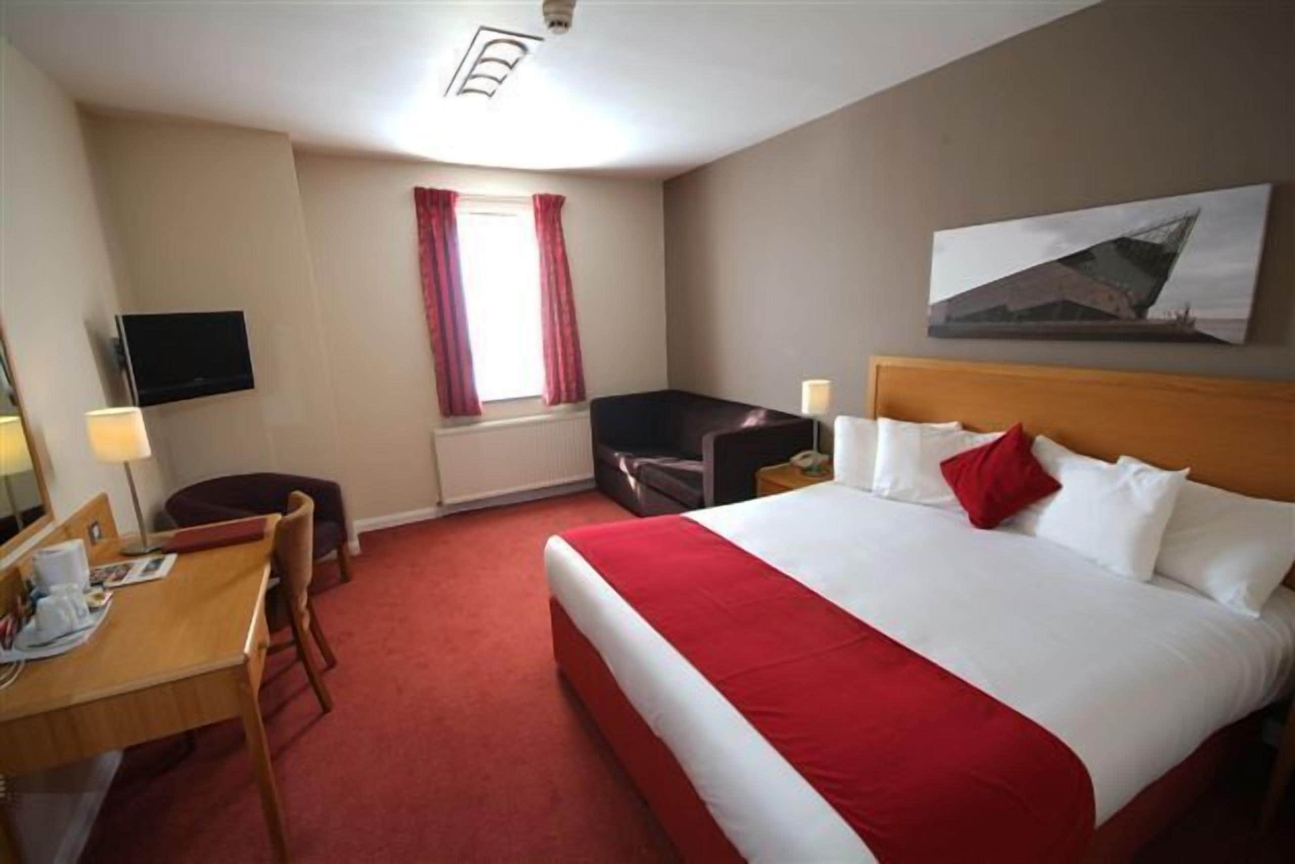 Una habitación de hotel con cama, escritorio, silla, televisión y ventana con cortinas.