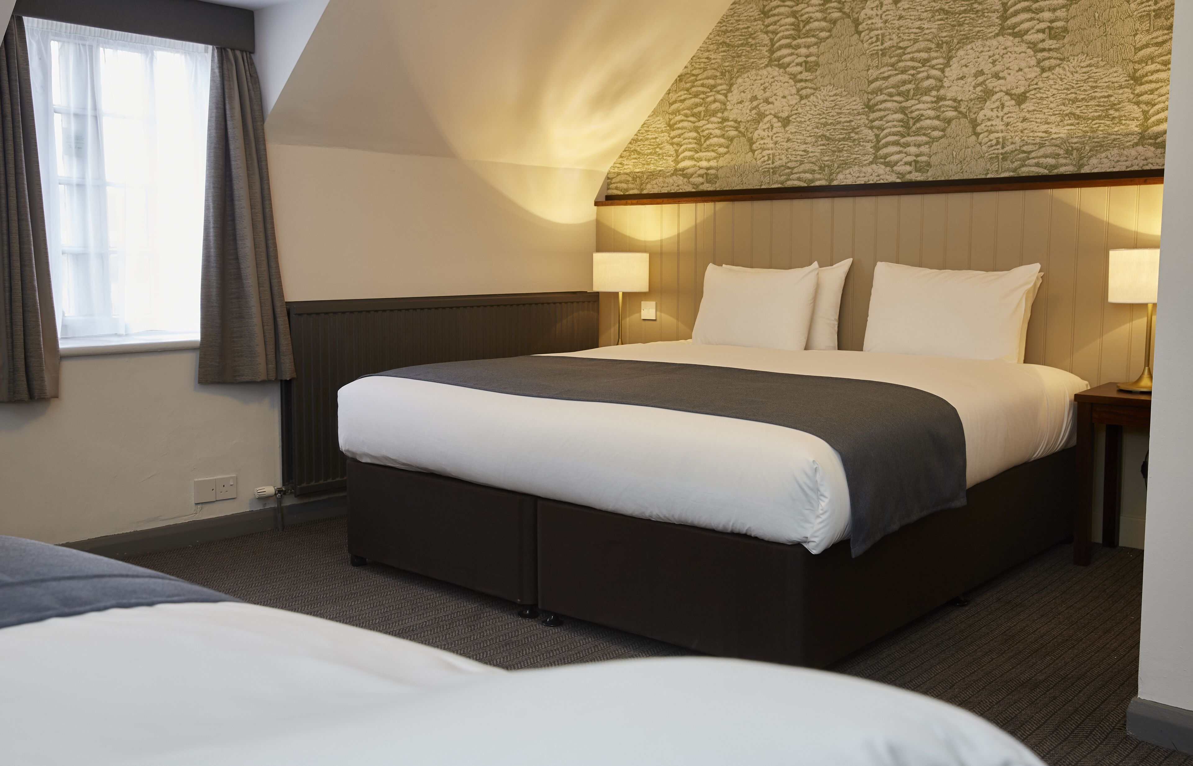Ein Hotelzimmer mit einem großen Bett, zwei Nachttischen mit Lampen, einem Fenster mit Vorhängen und einer gemusterten Decke.