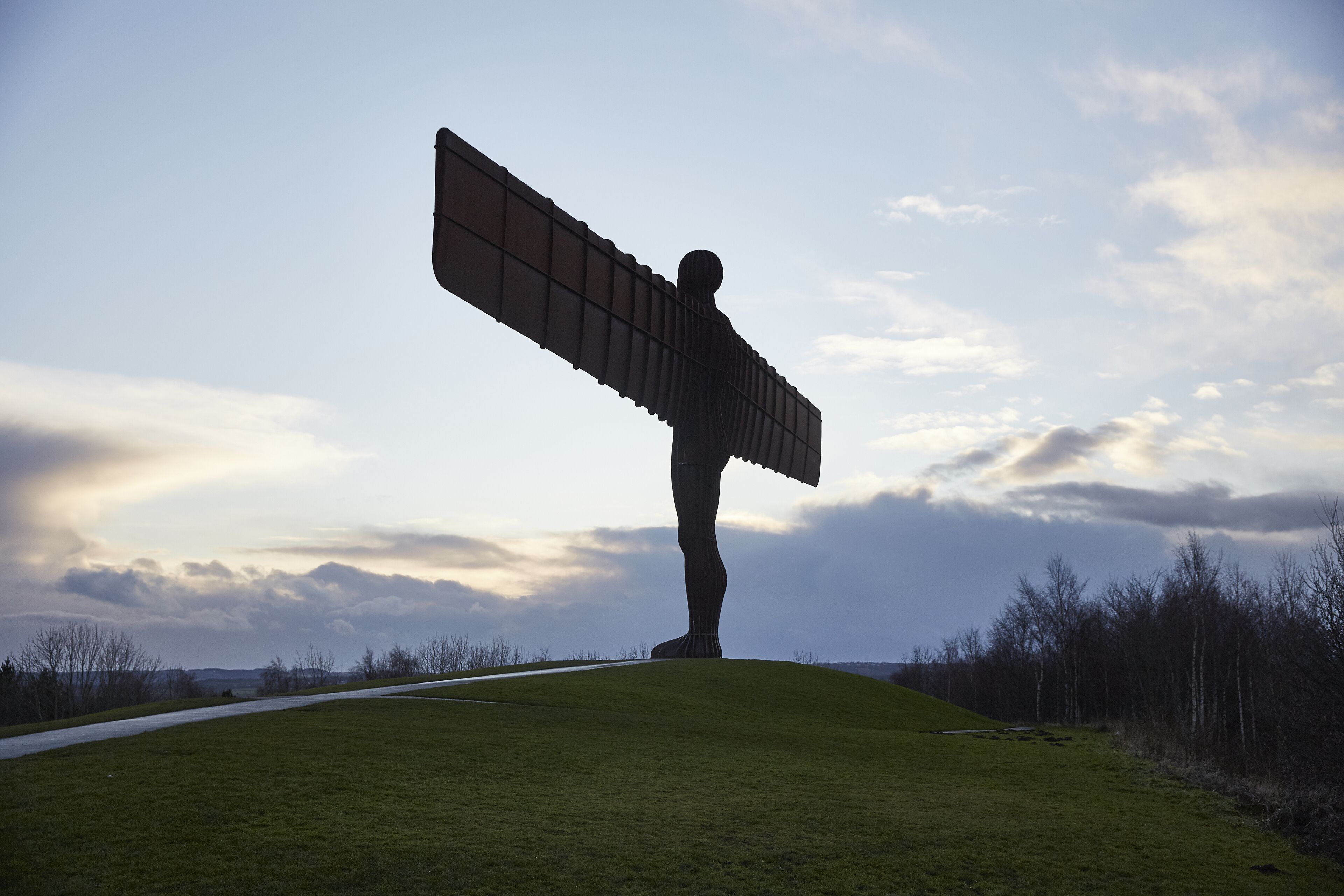 A large sculpture of a figure with outstretched arms on a grassy hill with a path and trees in the background.