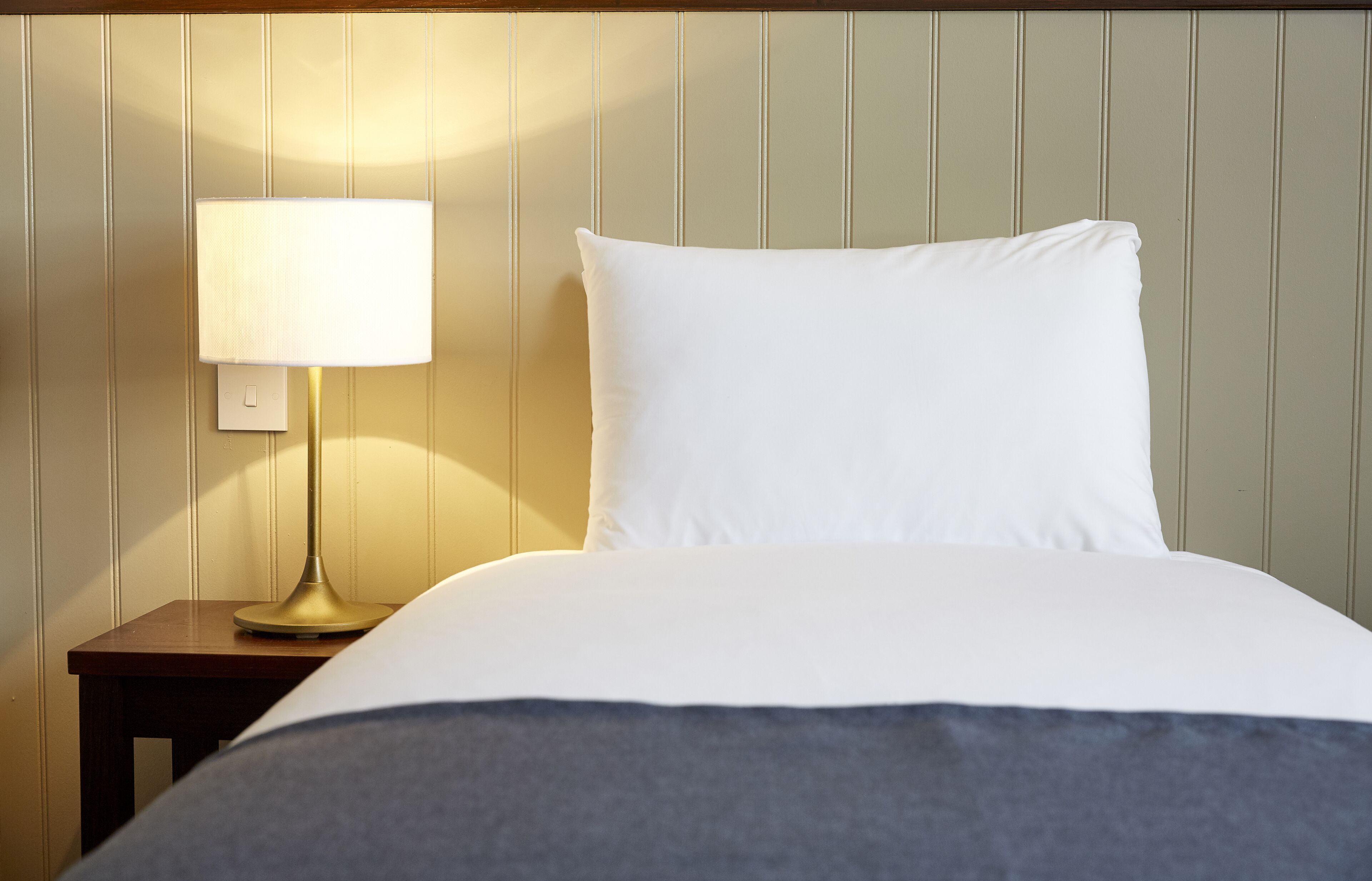 Een opgemaakt bed met witte kussens en een donkere deken, een bedlamp op een houten nachtkastje en een wand met verticale panelen.