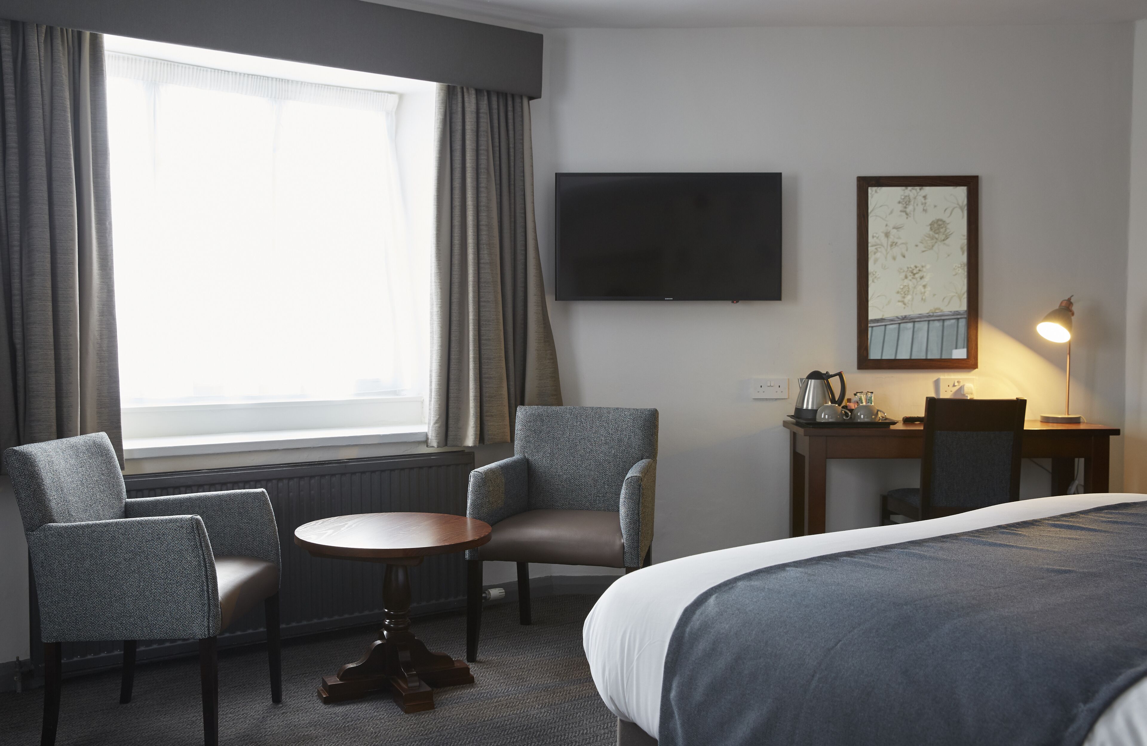 Habitación de hotel con cama, dos sillones, una mesita redonda, escritorio con lámpara, televisor de pantalla plana y ventana con cortinas.