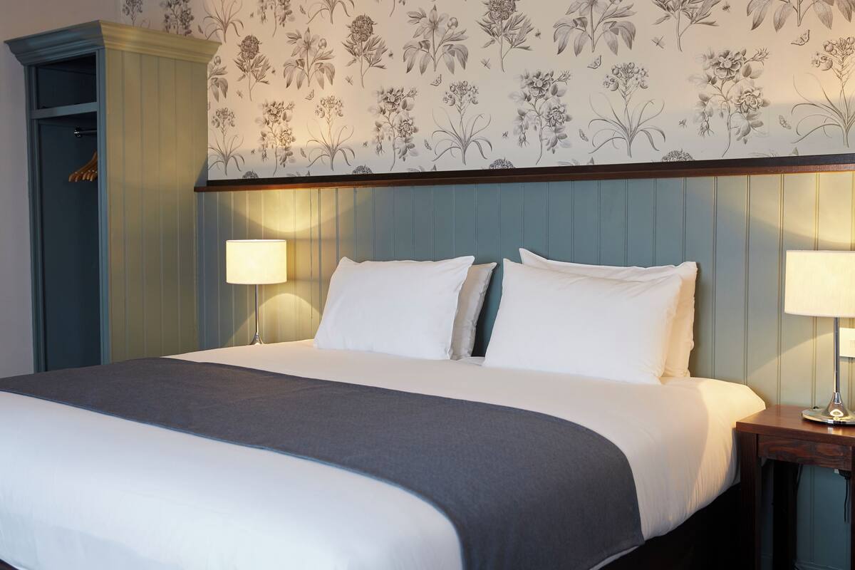 A neatly made bed with white and gray linens, a wooden nightstand with a lamp, and a floral wallpaper pattern on the wall.