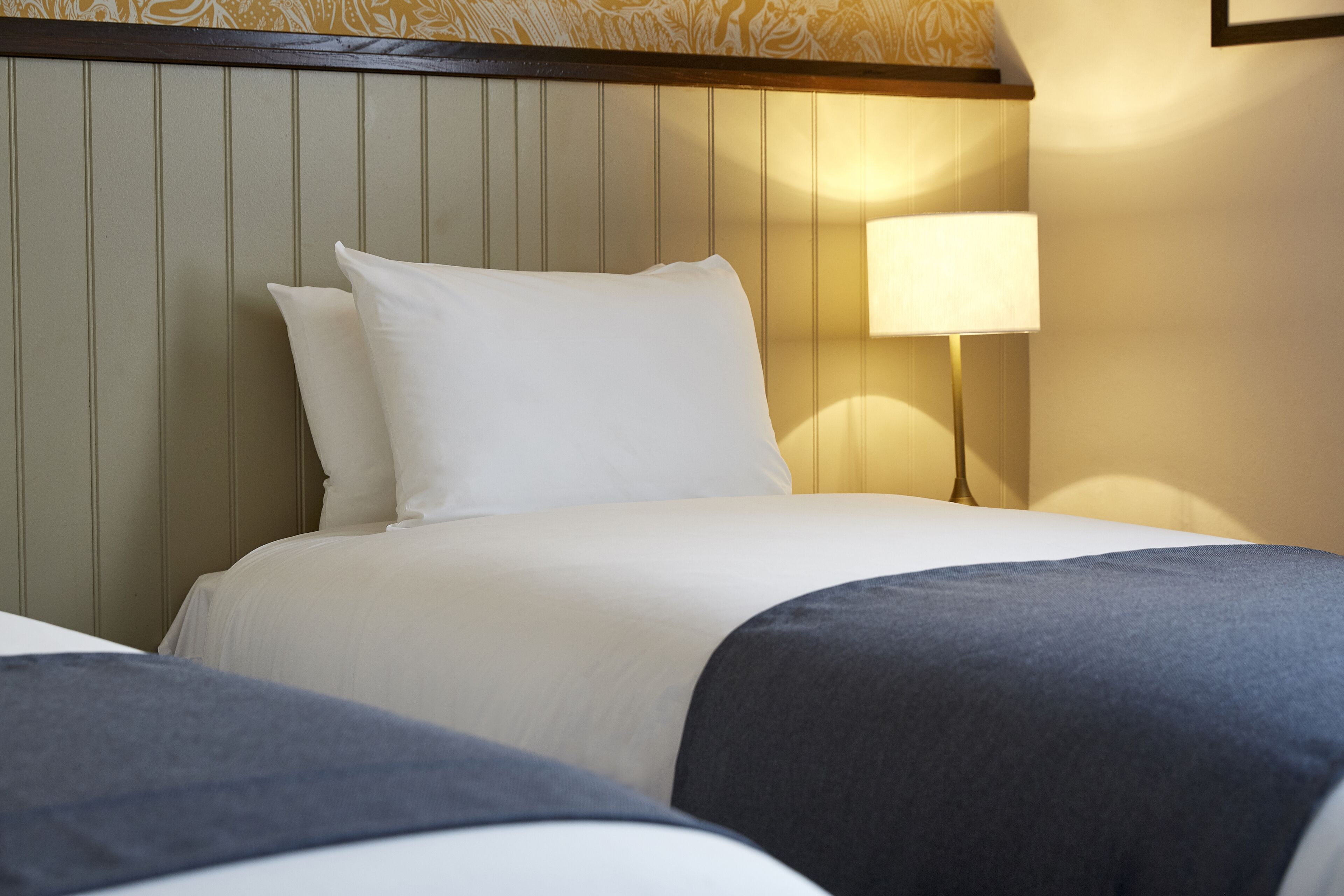 Una habitación de hotel con cama, almohadas, una lámpara de noche y cabecera con líneas verticales.
