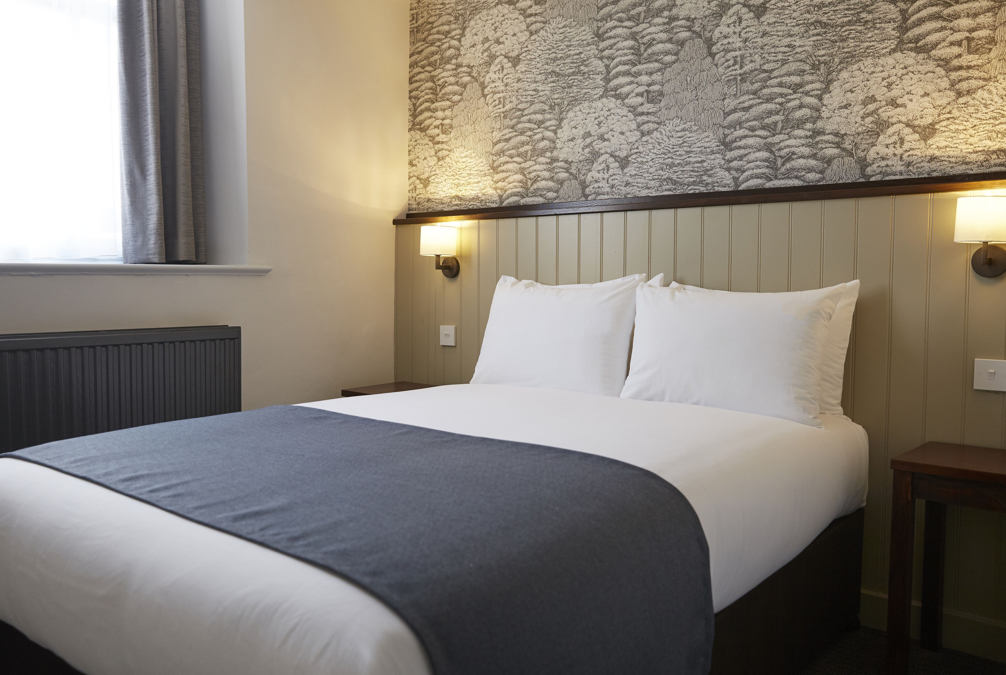 Una cama bien tendida con sábanas blancas y una manta gris, una lámpara de pared y una pared texturizada sobre la cama.