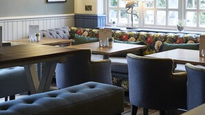 A cozy dining area with wooden tables and blue upholstered chairs, a bench with floral upholstery, and a large window with a view of trees.