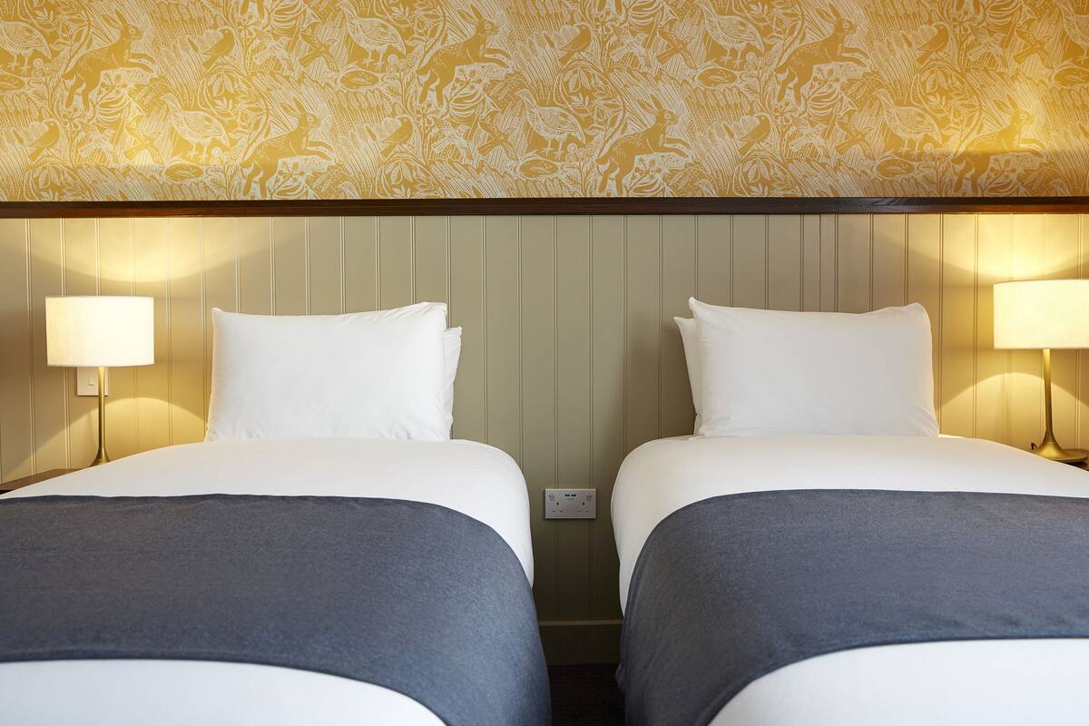 A hotel room with two beds, each with a white pillow and a grey blanket. There are wall-mounted lamps on either side of the beds.