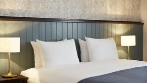 A neatly made bed with white linens and two bedside lamps, set against a wall with a floral pattern and a wooden headboard.