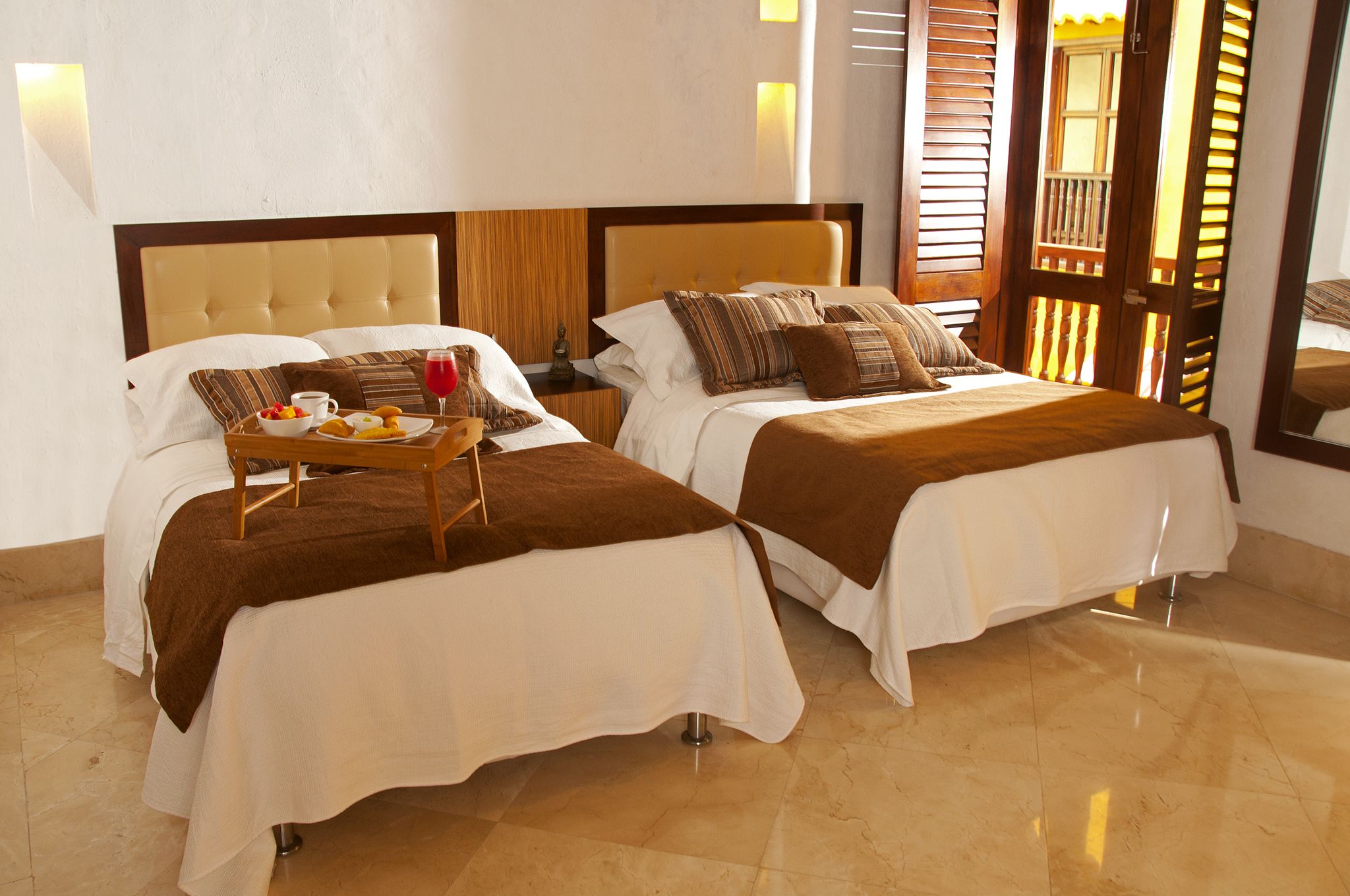 Habitación de hotel con dos camas, cada una con una manta y almohadas marrones, una bandeja con frutas y un vaso de bebida roja, y un cabecero de madera.