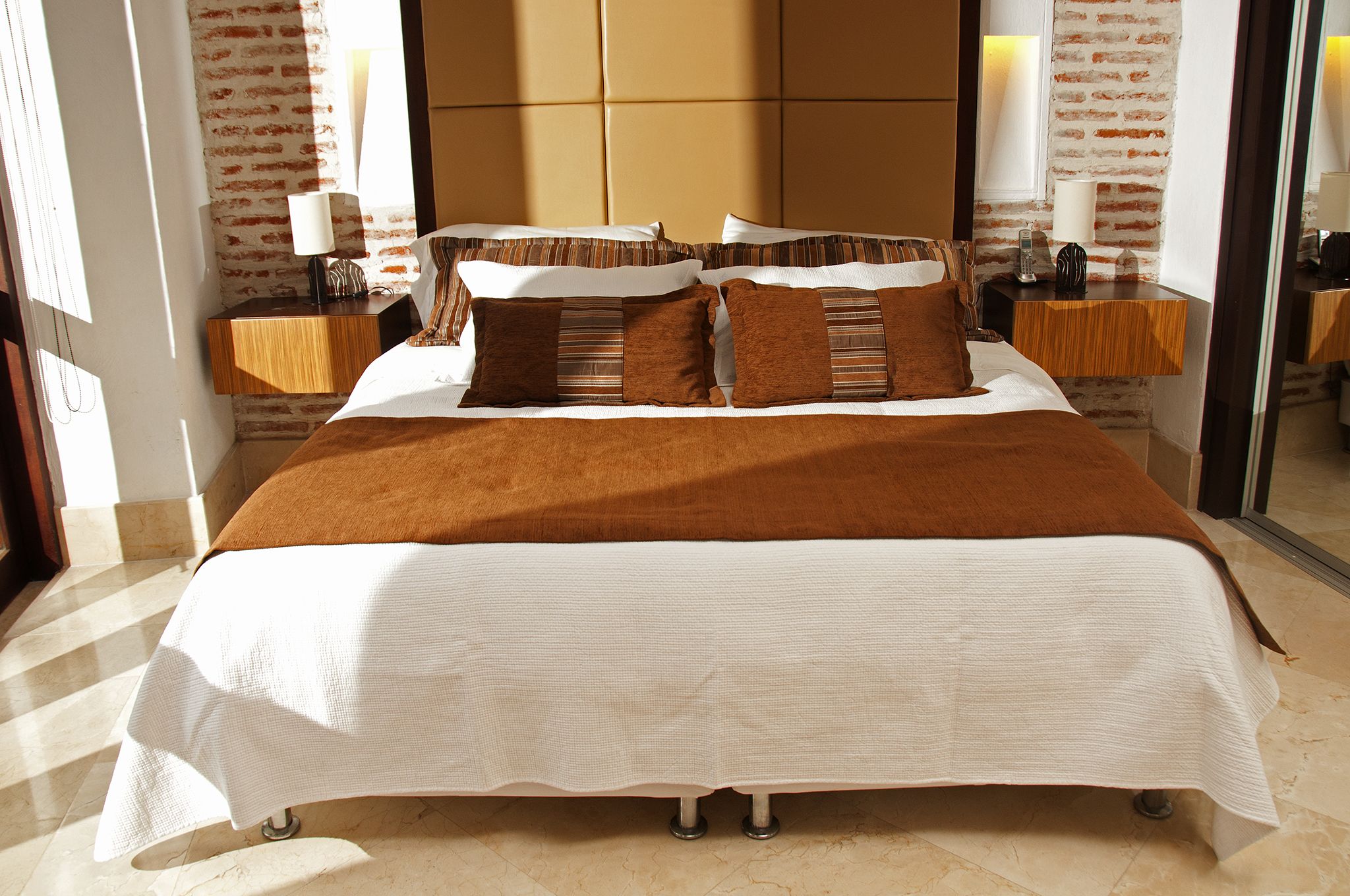 A neatly made bed with a brown blanket and white bedding, two bedside tables with lamps, and a brick-patterned wall.