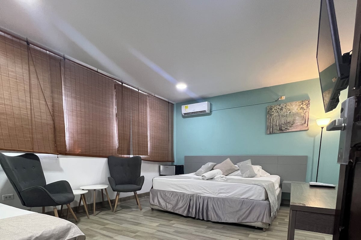 A hotel room with a bed, two chairs, a desk, an air conditioner, and a painting on the wall.