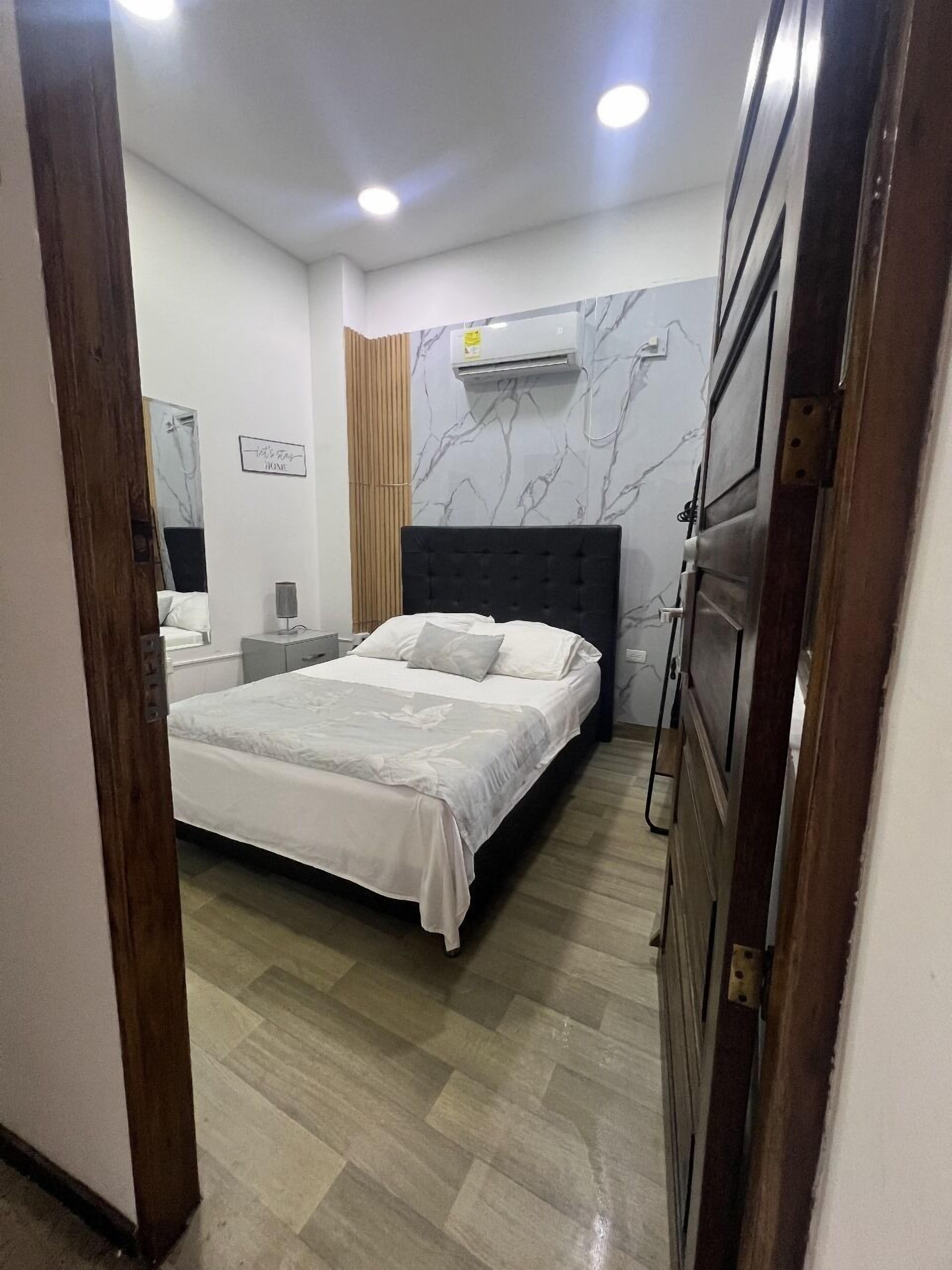 A bedroom with a bed, bedside table, lamp, air conditioning unit, and a mirror.