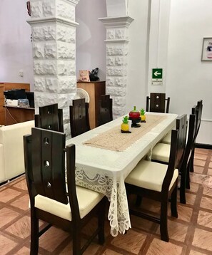 Un comedor con una mesa larga, mantel de encaje blanco y sillas de madera oscura. Se observan dos columnas blancas con diseños texturizados, un letrero de salida de emergencia verde y un estante con objetos.