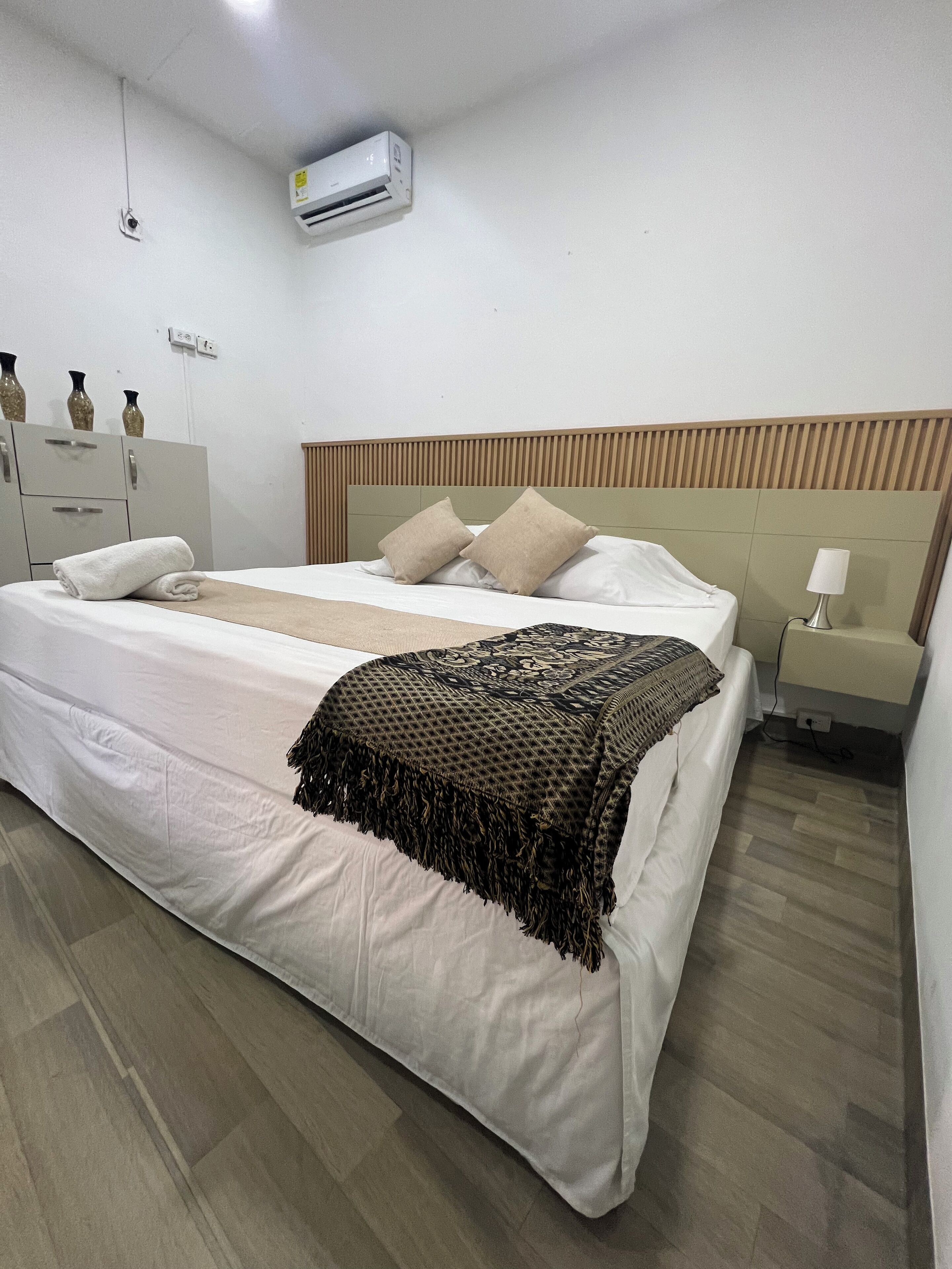 A hotel room with a bed, a nightstand, a lamp, an air conditioning unit, and a wardrobe.