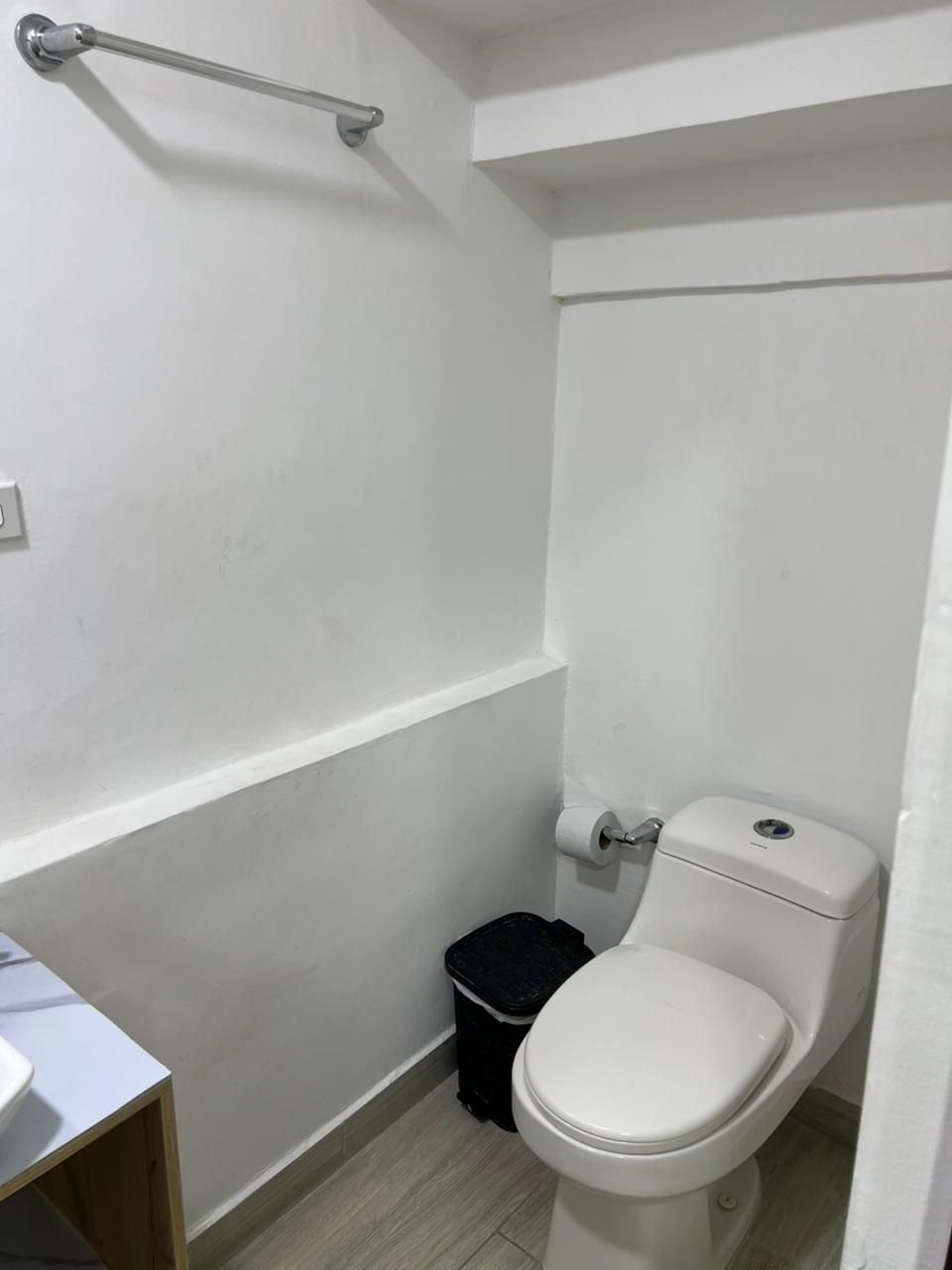 A small, white-walled bathroom with a white toilet, a black trash bin, and a wooden cabinet.