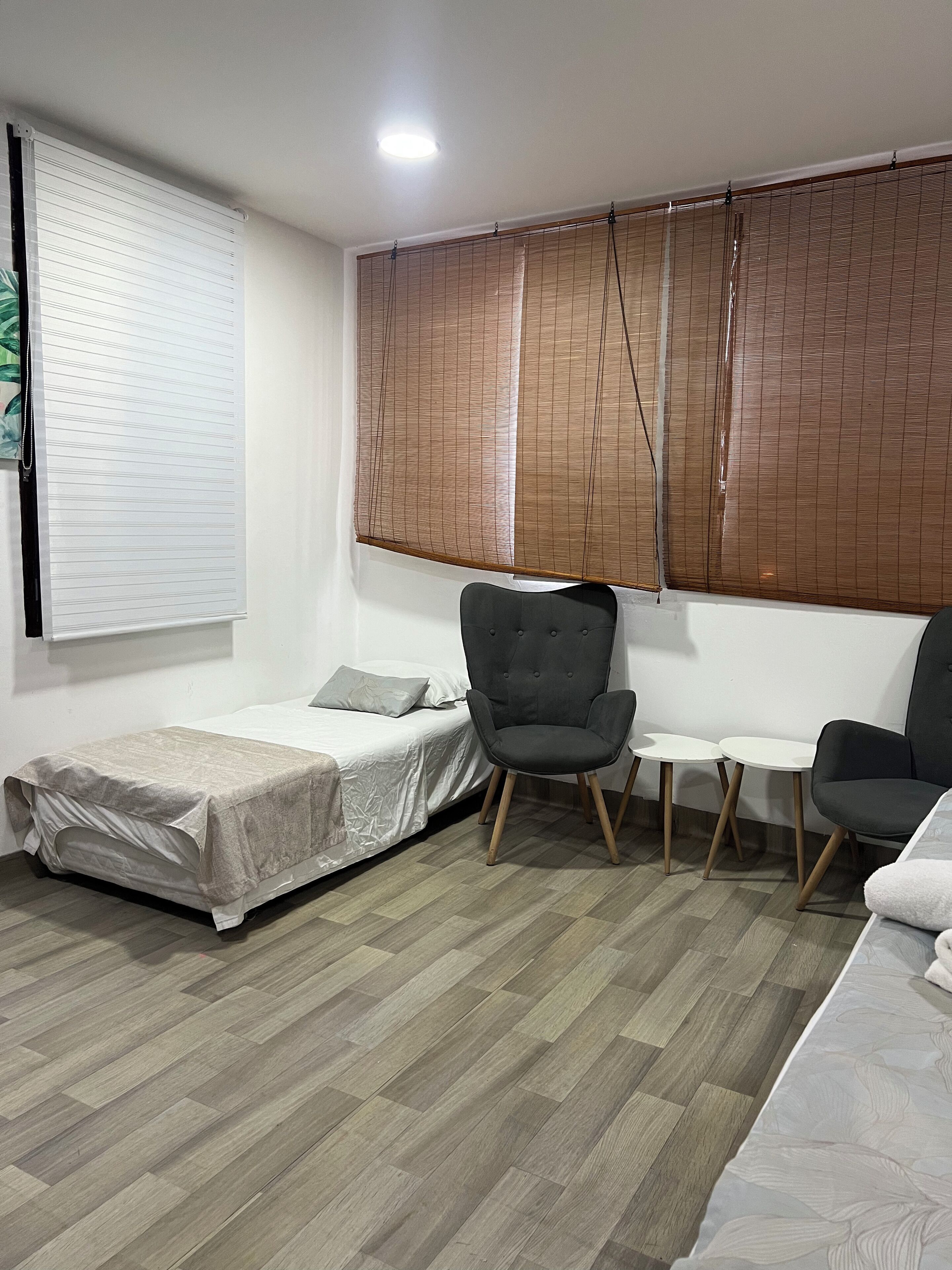 A small, modern room with a bed, two chairs, a small table, and a large window with blinds.