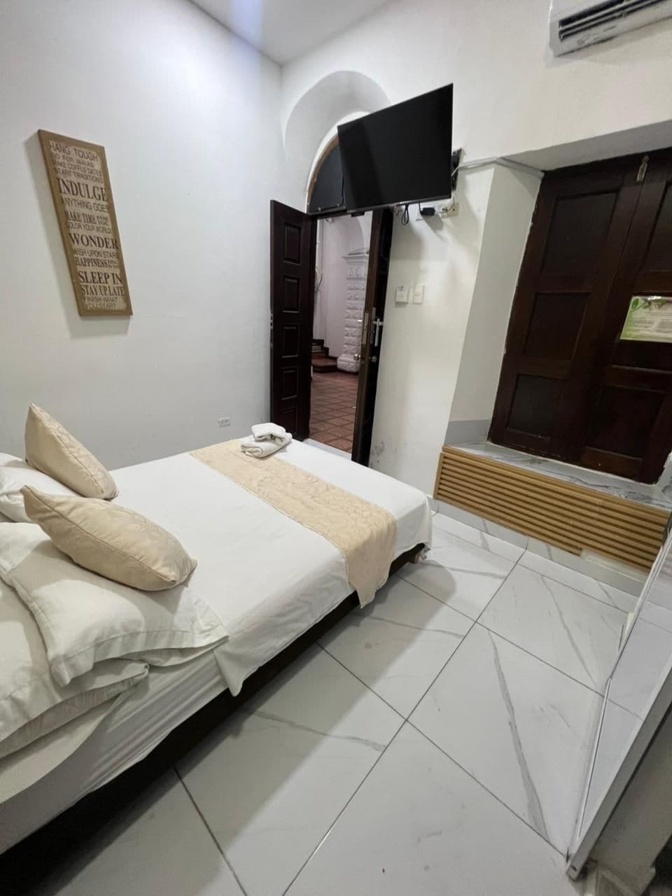 A bedroom with a bed, pillows, a flat-screen TV mounted on the wall, a wooden door, and a small window.