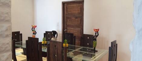 Un comedor con una mesa de vidrio rodeada de sillas de madera con asientos acolchados. Hay objetos decorativos sobre la mesa, como una jarra y pequeñas figuras.