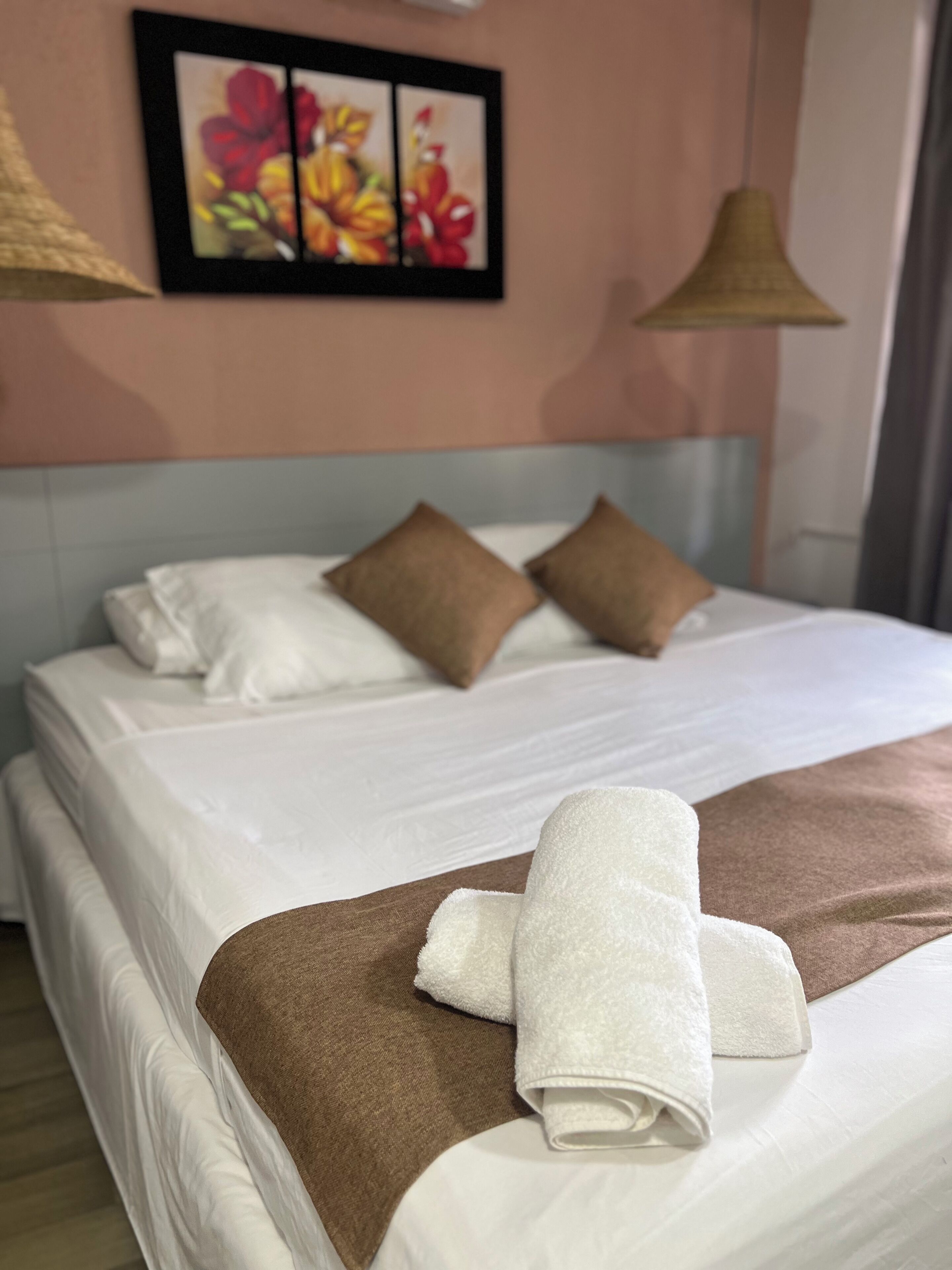 A neatly made bed with white and brown bedding, a framed floral artwork on the wall, and a hanging lamp.