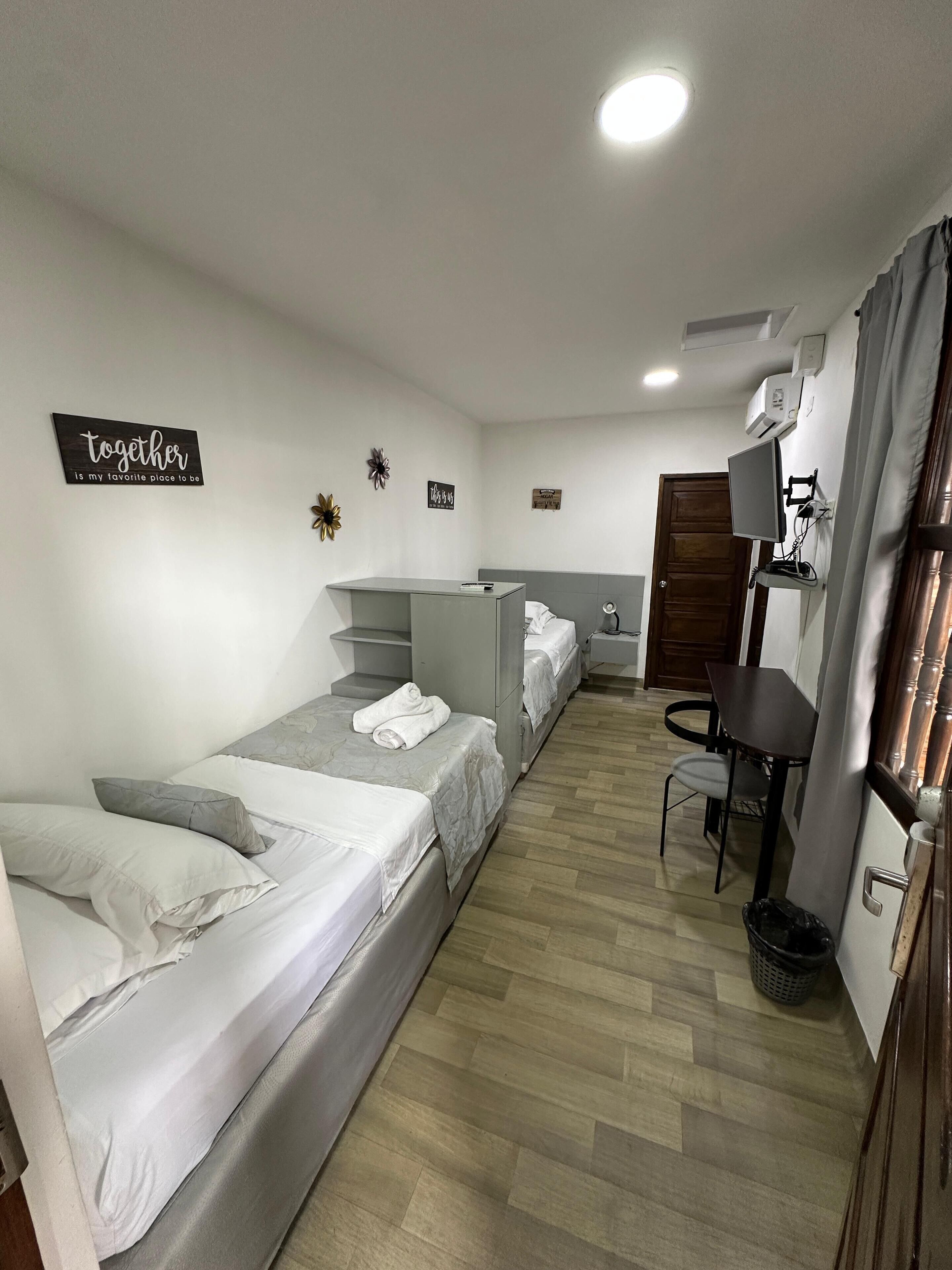Quarto em dormitório com duas camas individuais, uma escrivaninha, uma cadeira, uma mesinha e uma televisão.