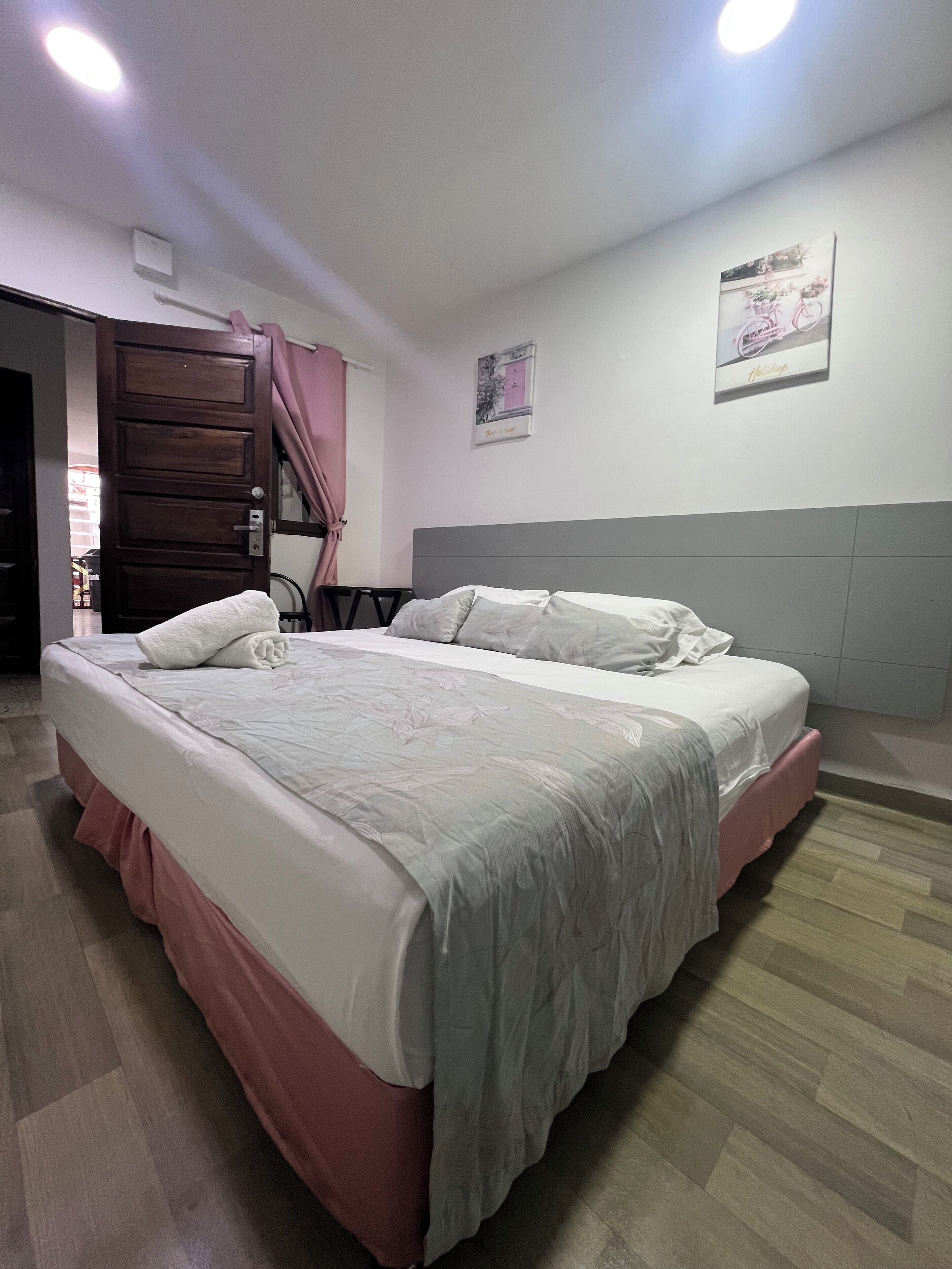 Ein ordentlich bezogenes Bett mit rosa Bettdecke, grauem Kopfteil und einer weißen Wand mit zwei gerahmten Bildern.