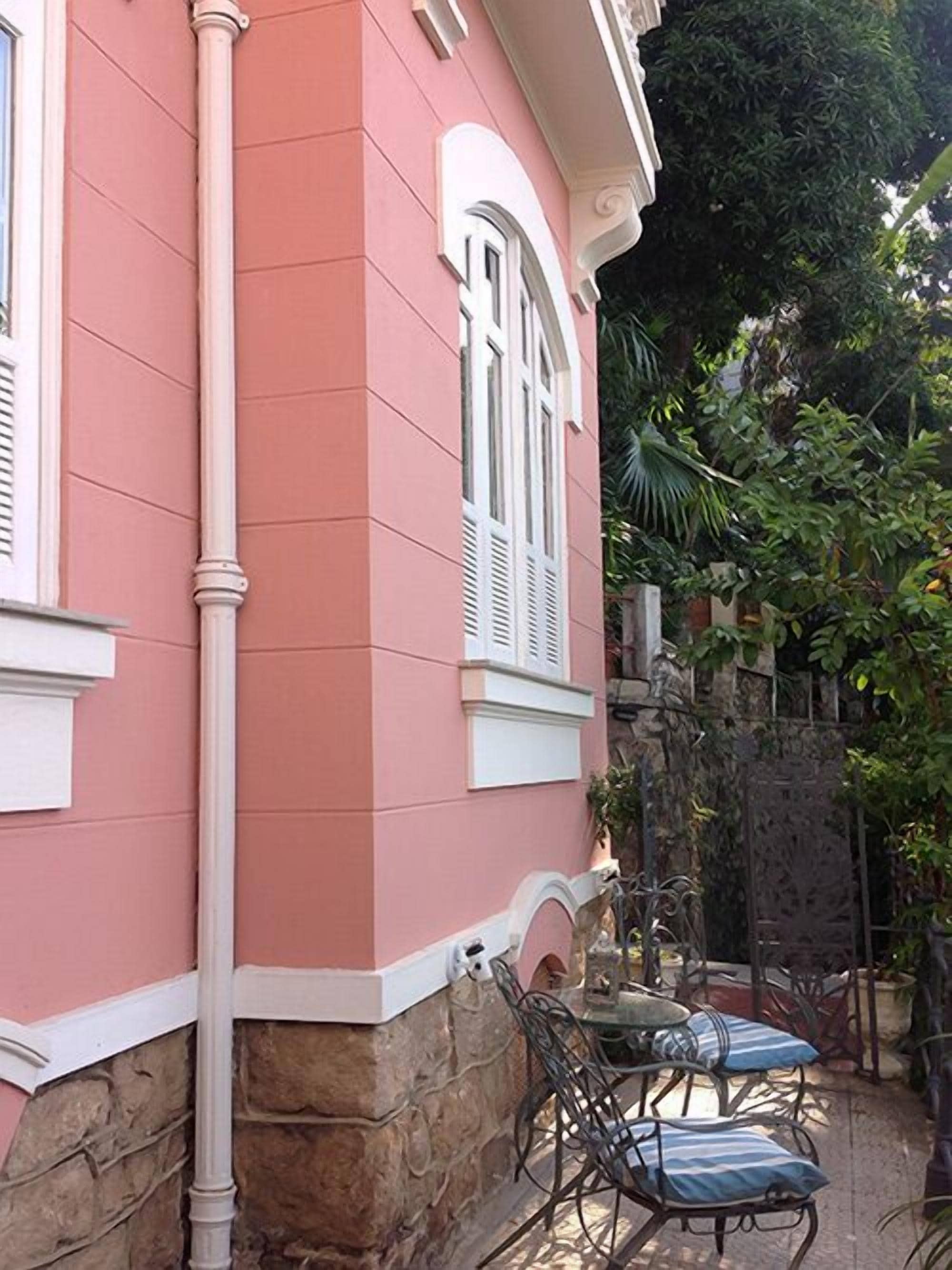 Un edificio rosa con detalles blancos, base de piedra y una ventana con contraventanas. Hay una mesa y dos sillas afuera.