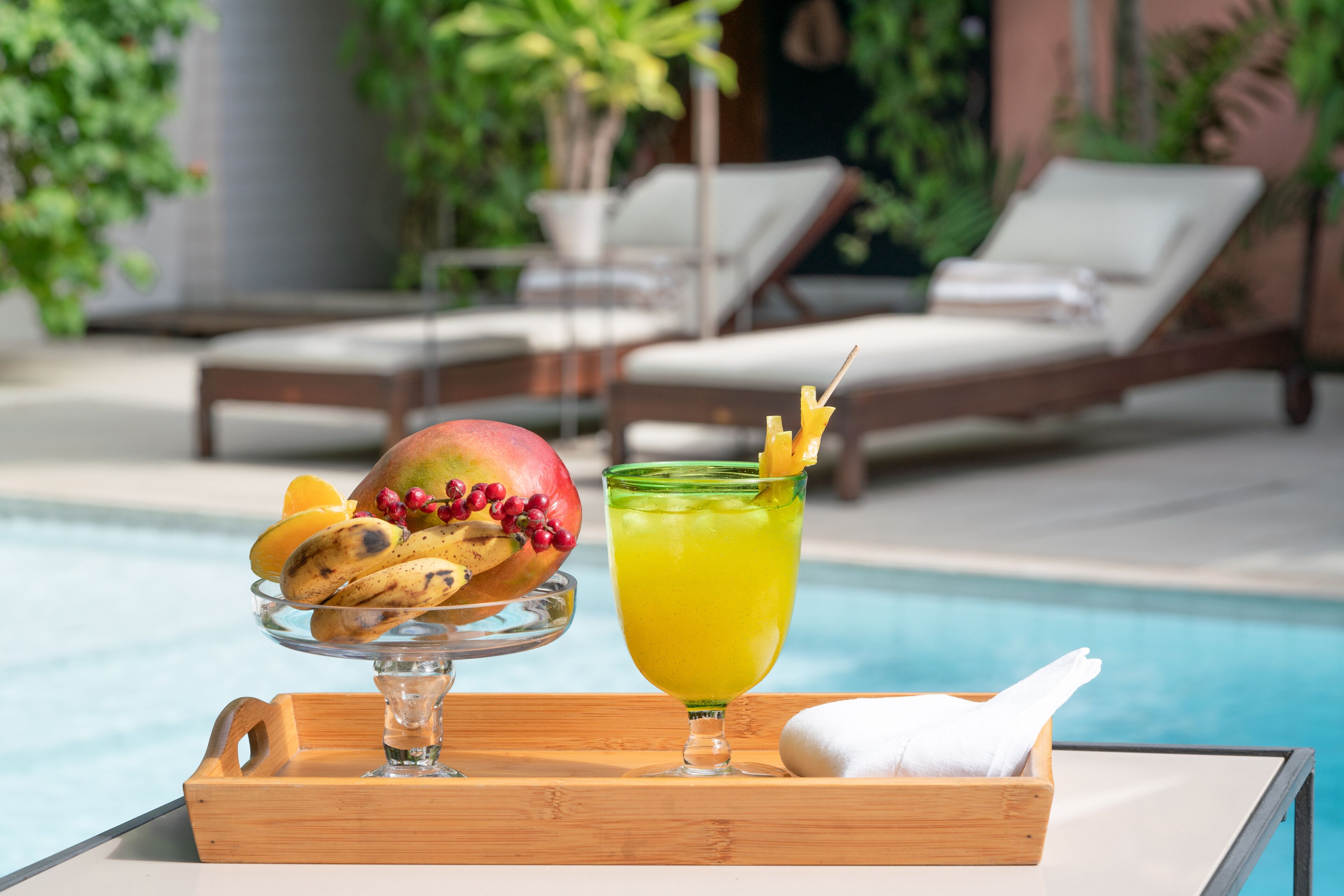 A tray with a fruit bowl and a glass of yellow drink, a napkin, and a poolside setting with lounge chairs.