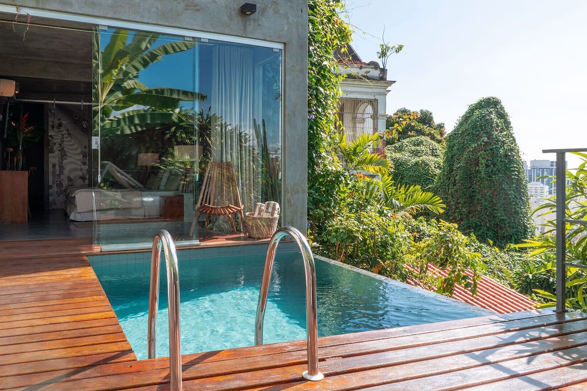 Ein moderner Poolbereich mit Holzterrasse, einem wintergartenartigen Raum mit Sofa und Sessel sowie üppiger Bepflanzung rund um das Anwesen.