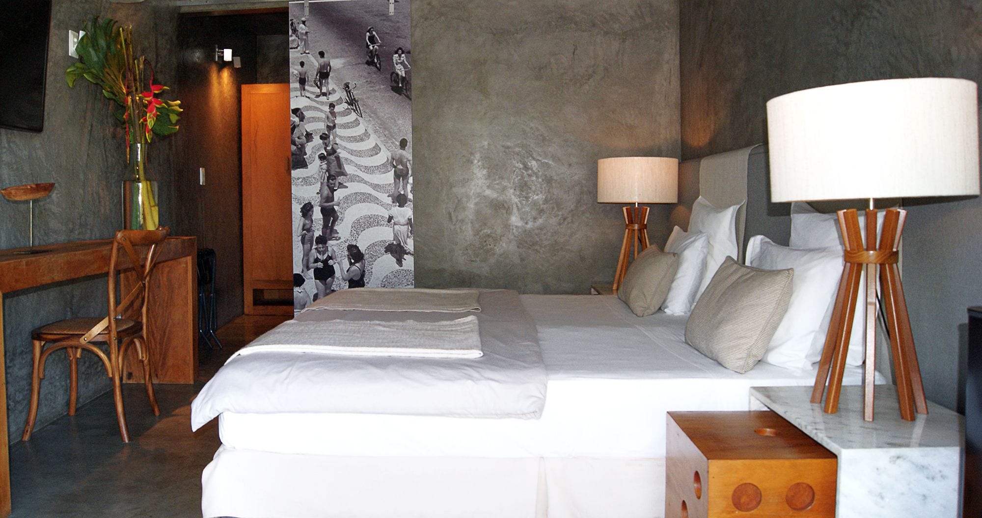 A hotel room with a bed, a desk, a chair, a lamp, and a wall with a patterned mural.