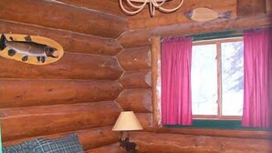 A log cabin bedroom with a bed, a lamp, a fish mounted on the wall, and a window with curtains. - Scandanavian Vacation Rental (Lake City)