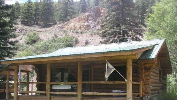 A log cabin with a porch, surrounded by a grassy area and dense forest. - Scandanavian Vacation Rental (Lake City)