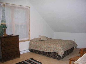 A bedroom with a bed, a dresser, a chair, and a window with sheer curtains.