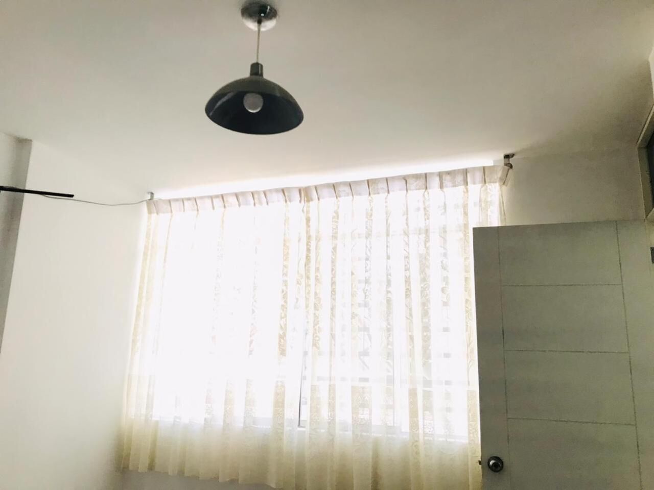Una habitación con ventilador de techo, ventana con cortinas de gasa y puerta con picaporte metálico.
