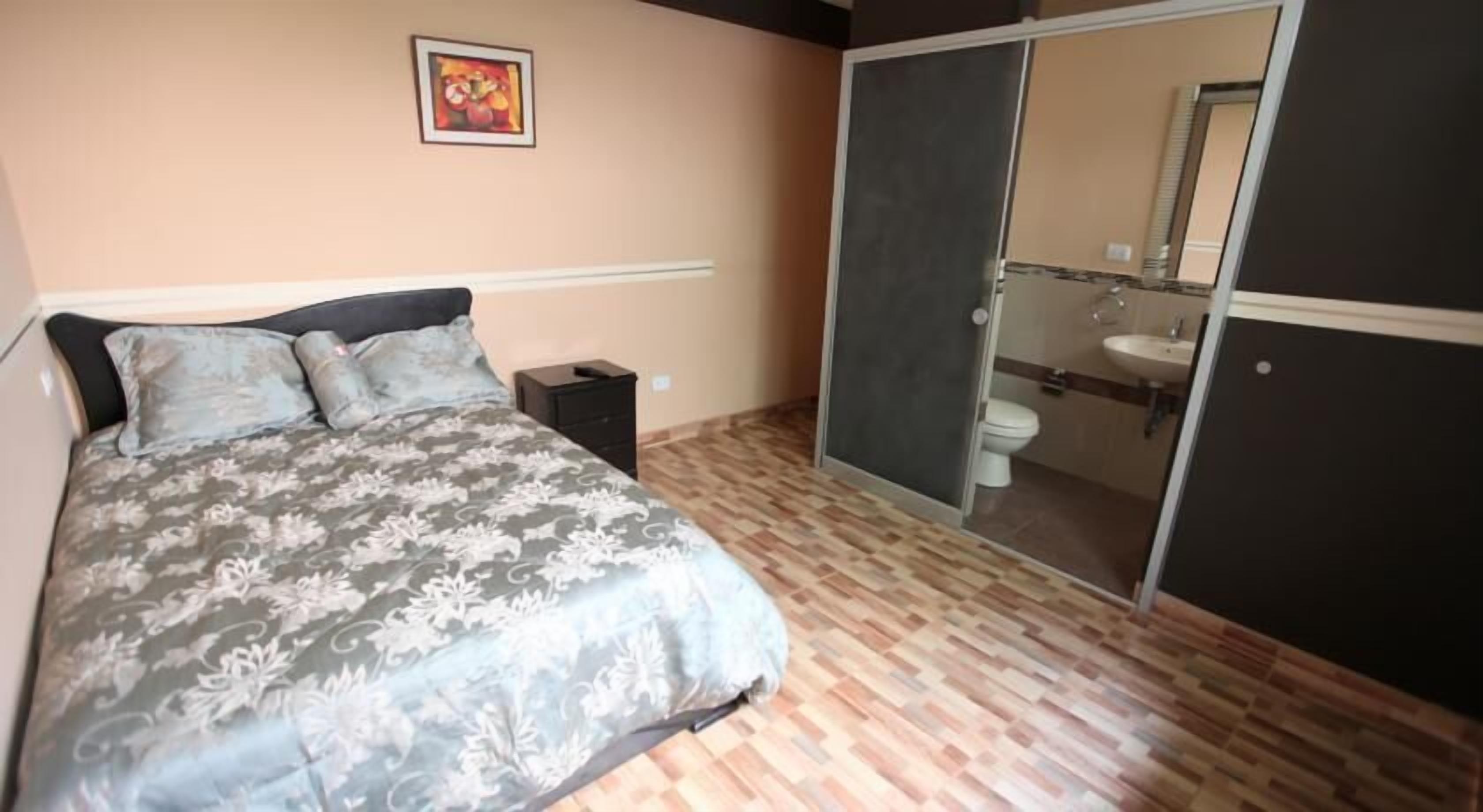 Un dormitorio con cama, mesita de noche, puerta corrediza al baño y un cuadro en la pared.