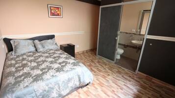 Un dormitorio con cama, mesita de noche, puerta corrediza al baño y un cuadro en la pared.