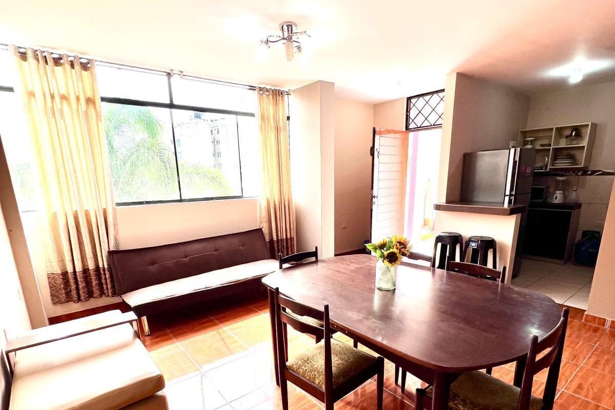 A dining area with a wooden table and chairs, a sofa, and a kitchen in the background.
