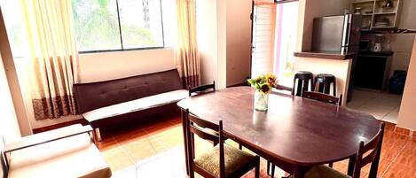 Uma sala de jantar com mesa e cadeiras de madeira, um sofá e uma cozinha ao fundo.