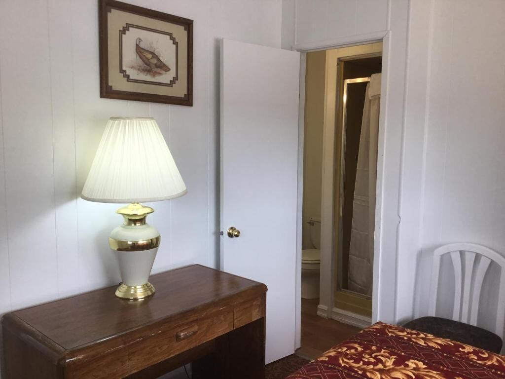 Une pièce avec un bureau en bois, une lampe, un tableau encadré au mur et une porte donnant sur une salle de bain.