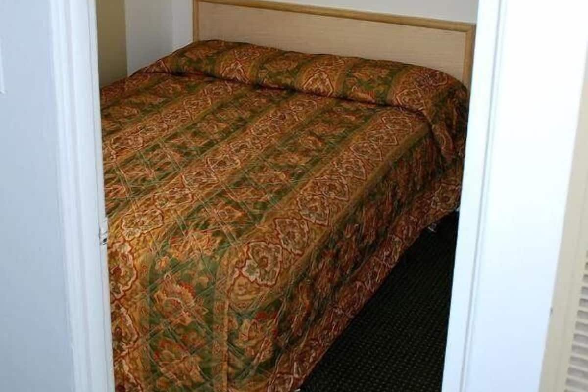 A single bed with a patterned bedspread in a room with a plain wall and a framed picture above it.
